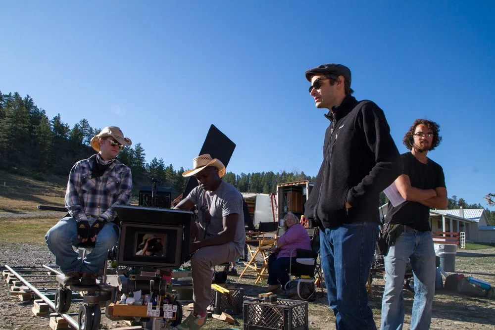 Jason Corgan Brown and crew on a film set outdoors with a camera, equipment, and trail tracks, under a clear blue sky, with trees and buildings in the background.