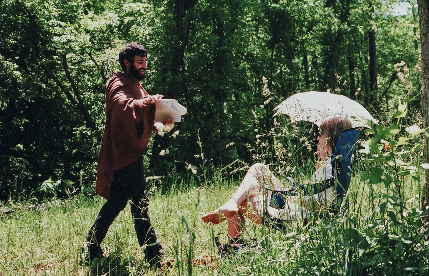 Creighton Barrett of Band of Horses dressed in a brown cloak is handing a white object to a woman sitting in a blue chair under a white lace umbrella in a wooded area with sunlight filtering through the trees. Directed by Jason Corgan Brown. 