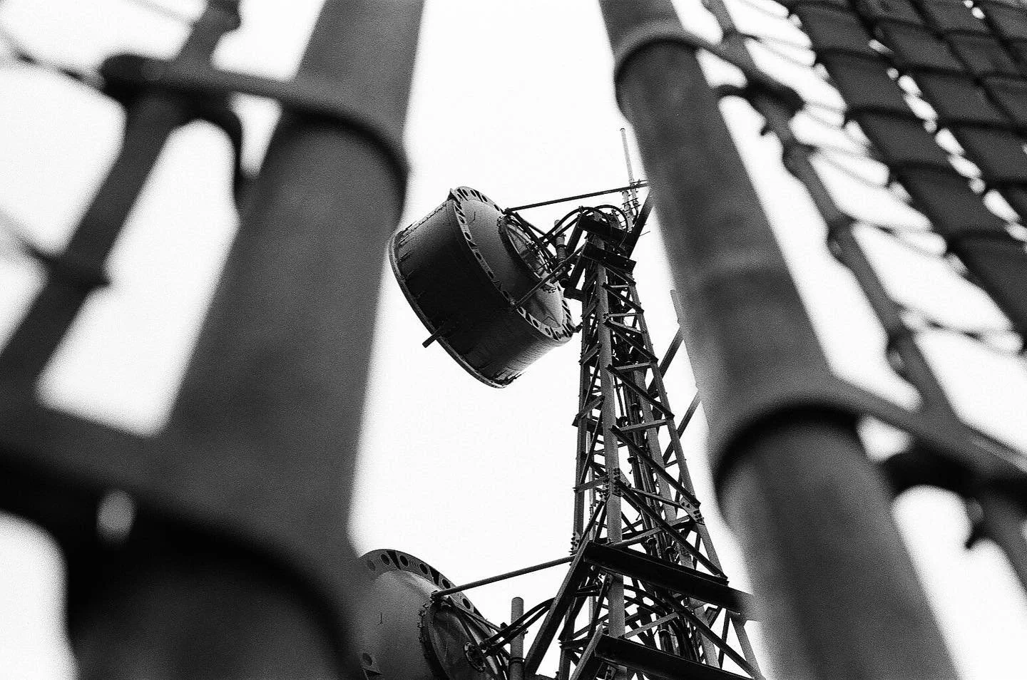 A black and white image of a tall industrial radio tower seen through a fence, with the camera angle pointing upward. Film photography by Jason Corgan Brown. 
