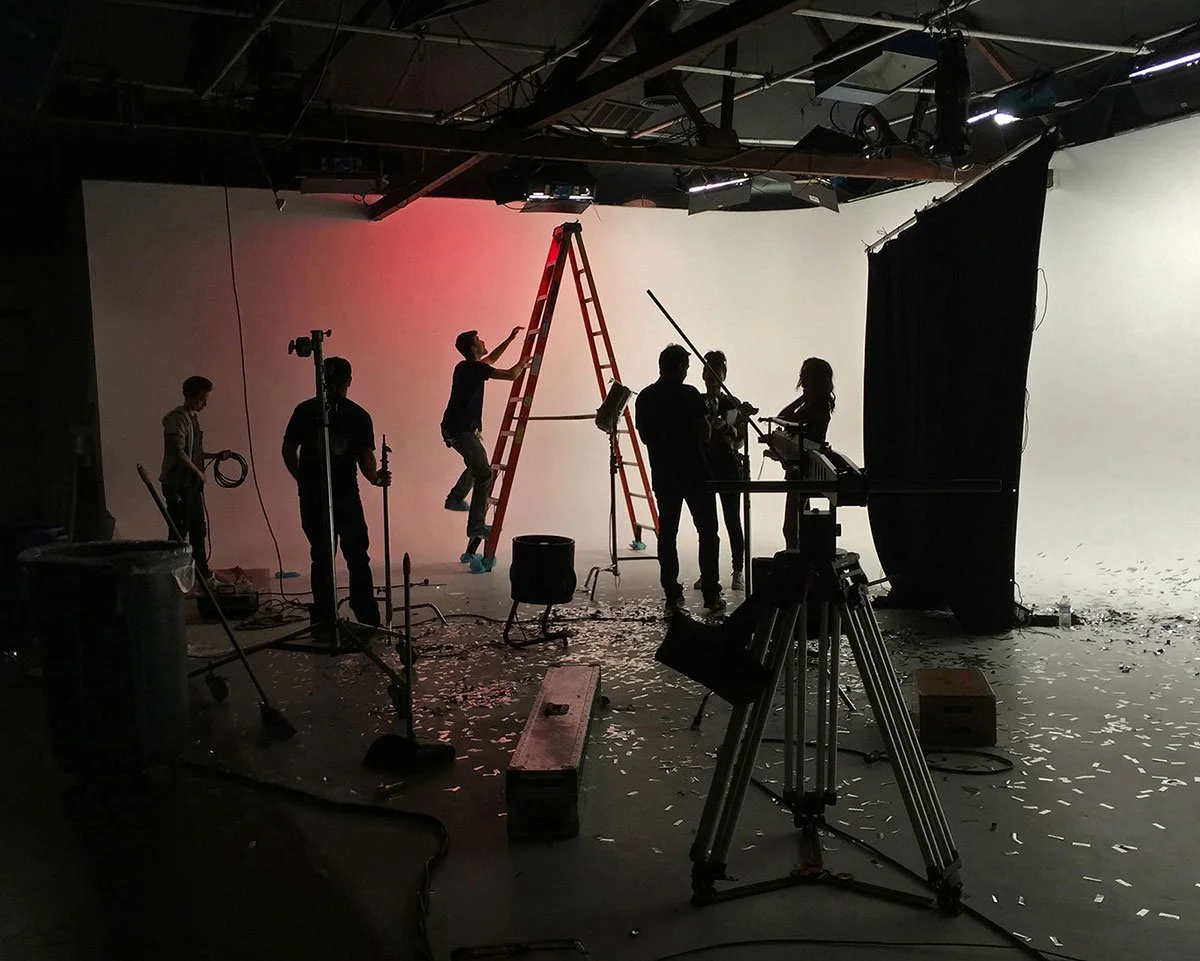 Corgan Studio film crew working in a studio with a white backdrop, using a ladder, cameras, and lighting equipment, with confetti on the floor. Directed by Jason Corgan Brown. 