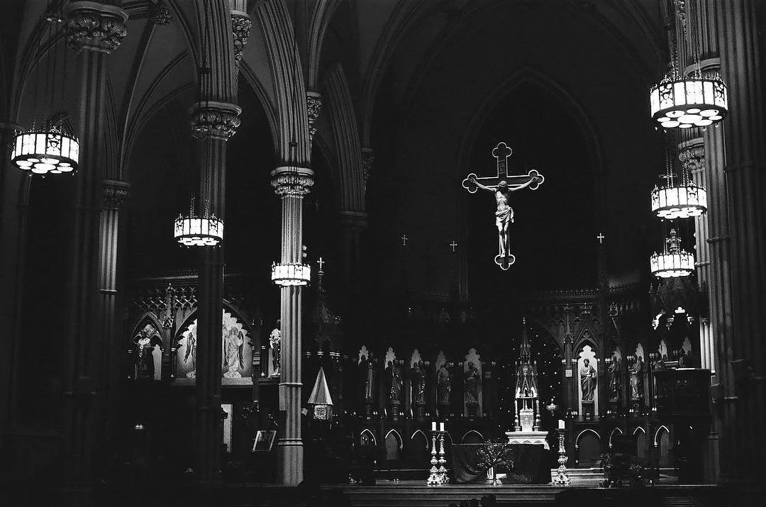 Interior of a Christian church with a large crucifix hanging above the altar, ornate chandeliers, and religious statues and artwork. Black and white film photography by Jason Corgan Brown. 