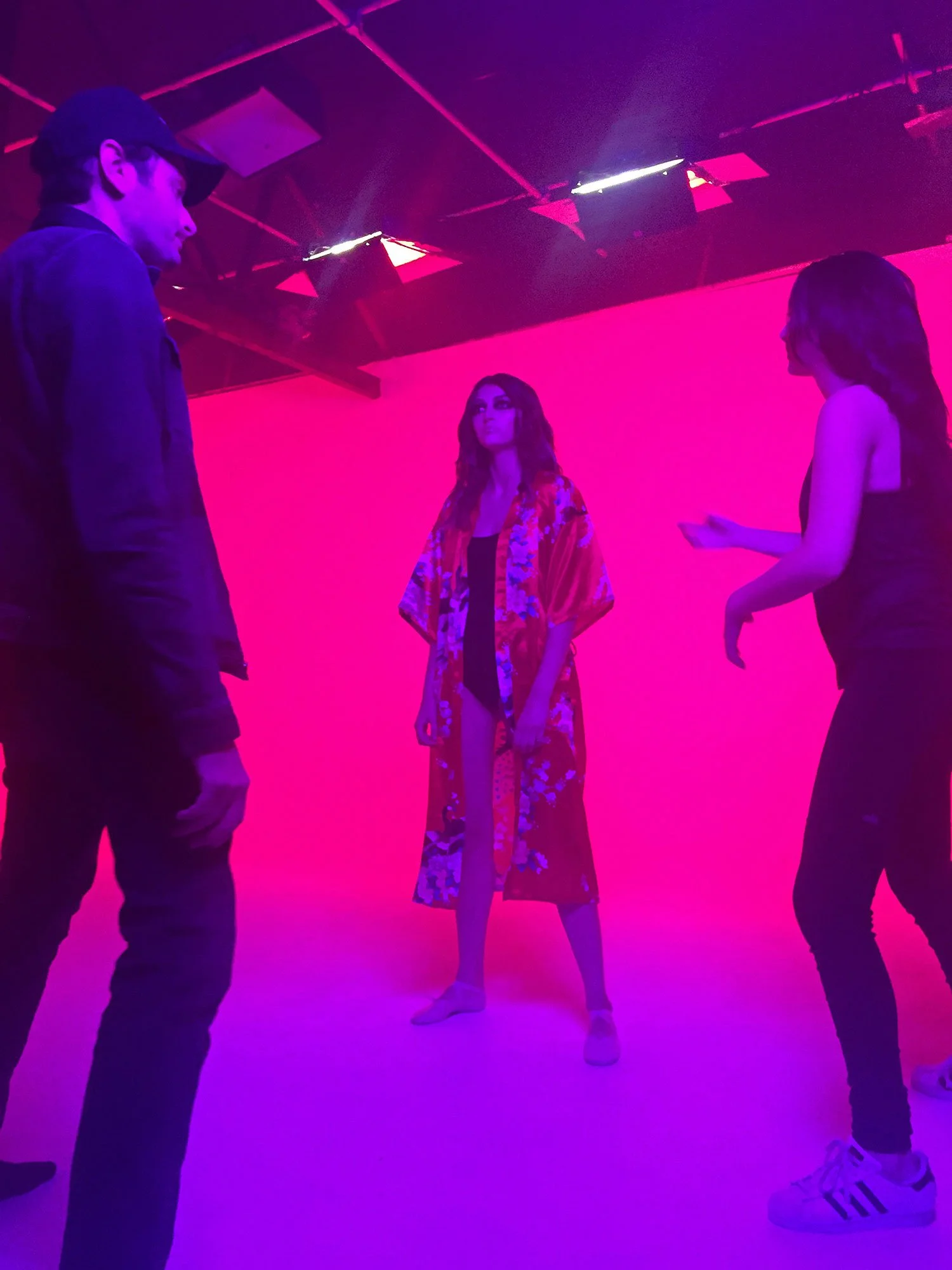 Jason Corgan Brown, Sasha Glasser, and Isabella Brenza in a neon pink and purple light studio, with a woman in the center wearing a floral kimono and the other two in dark outfits, engaged in conversation or coaching.