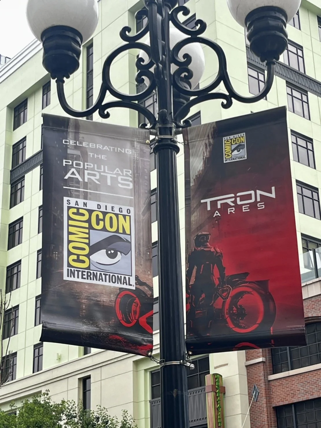Two banners on a streetlamp, one celebrating San Diego Comic-Con International and the other promoting the movie 'Tron: Ares' with a silhouette of a person and a motorcycle, against a city background.