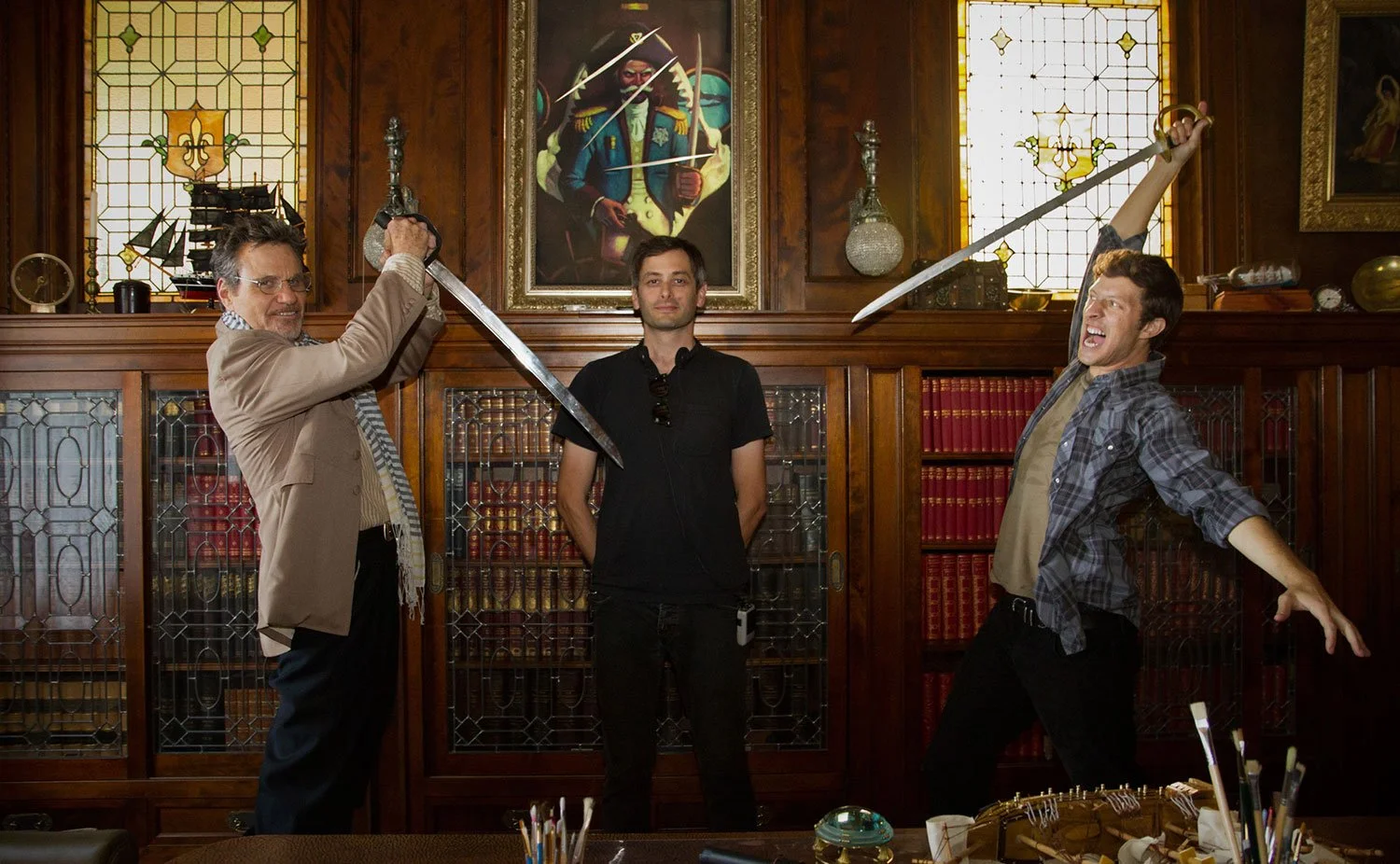 Jason Corgan Brown, Martin Kove, and Gabriel Sunday posing with swords on film set in a room with wooden bookshelves, stained glass windows, and framed paintings on the wall.