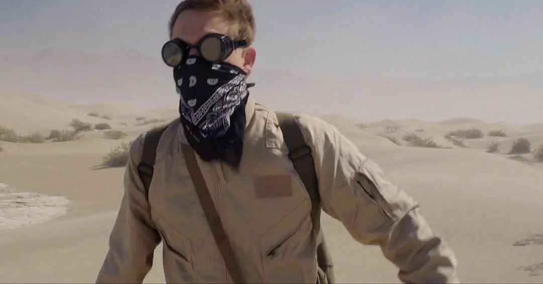 Jeter Rhodes wearing goggles, a bandana covering the lower face, a beige jacket, and a backpack walking in a desert with sand dunes. On a film set for Corgan Pictures, Directed by Jason Corgan Brown. 