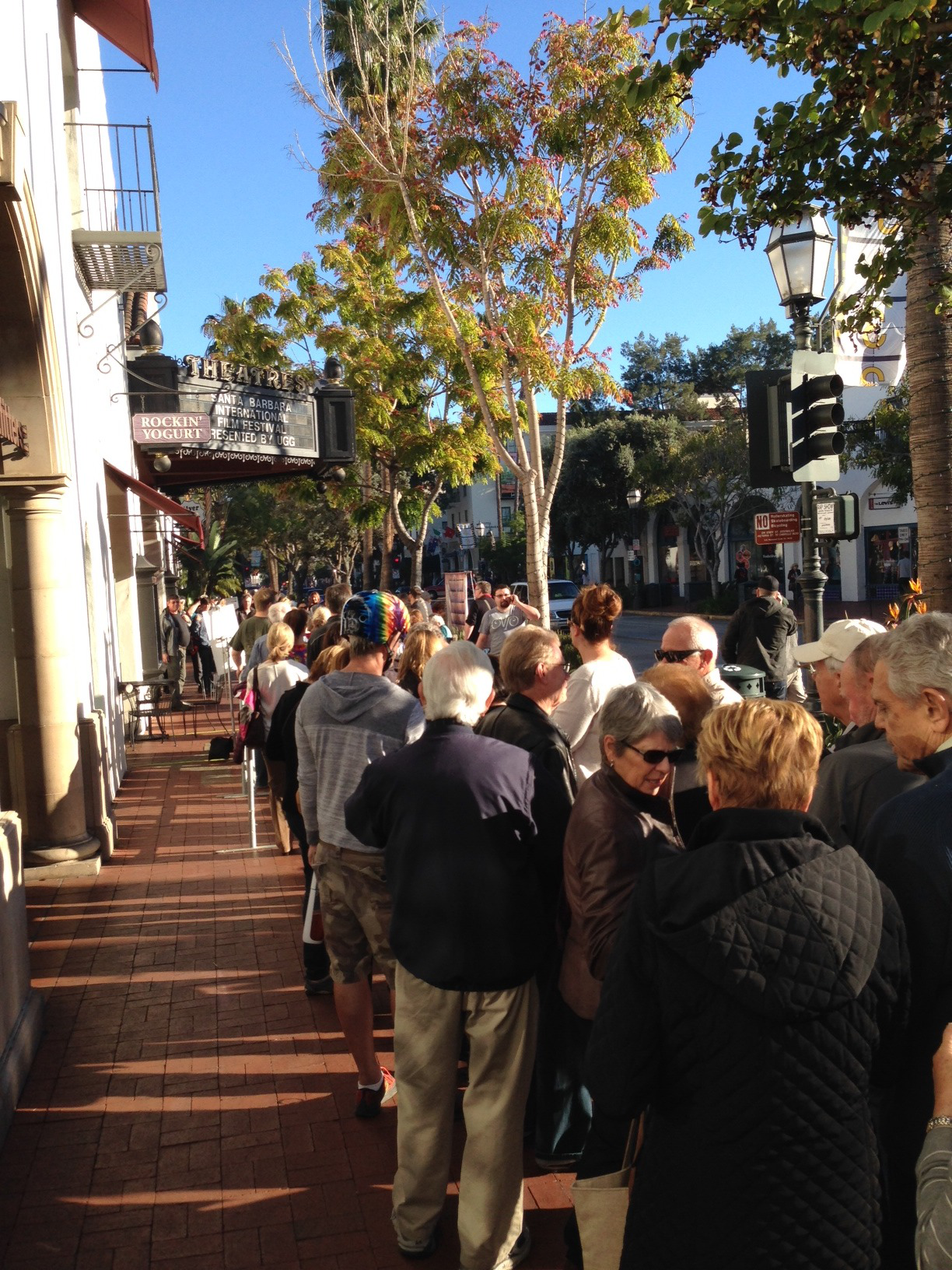 Long line of people waiting outside a store on a sidewalk in a shopping district, with trees, buildings, and a traffic light in the background.