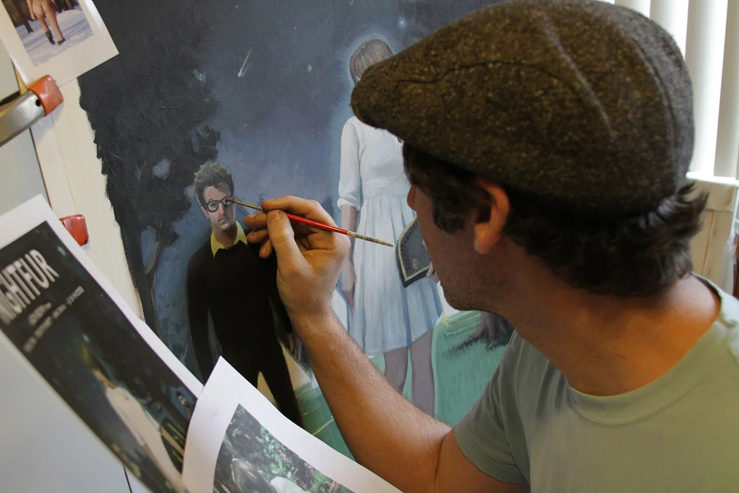 Matt Groller is painting a realistic portrait of a man with glasses on a canvas. The artist is wearing a gray cap and a light gray shirt, working with a fine brush in a well-lit room. There are photographs and images pinned on the wall nearby.