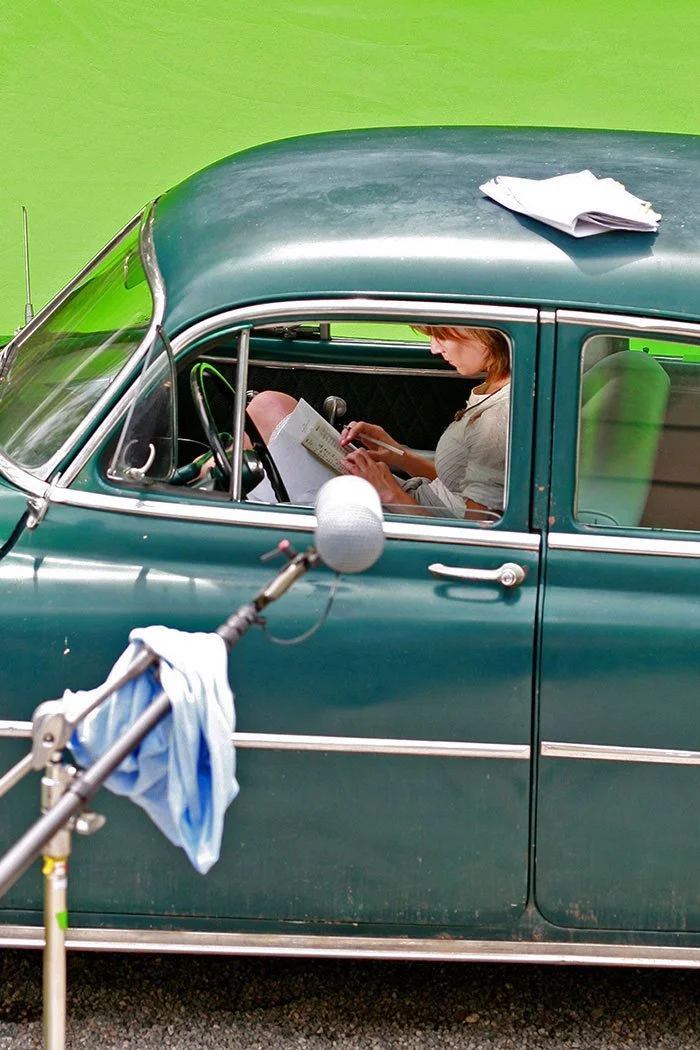 A woman sitting inside a vintage green car, reading a book or script, with a piece of paper on the roof, set against a solid green background, during a film shoot. Directed by Jason Corgan Brown. Starring Creighton Barrett of Band of Horses. 