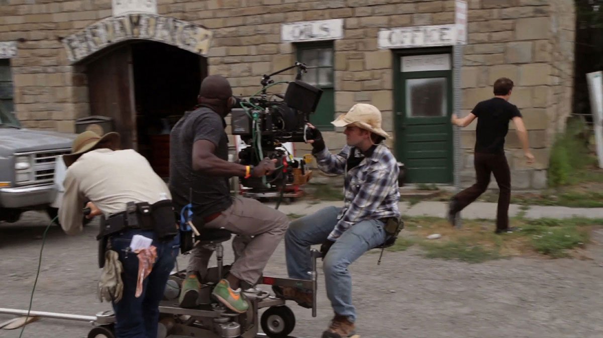 Corgan Studio crew members filming movie scene outside a stone building with signs saying 'Tavern & Lunch' and 'Café'