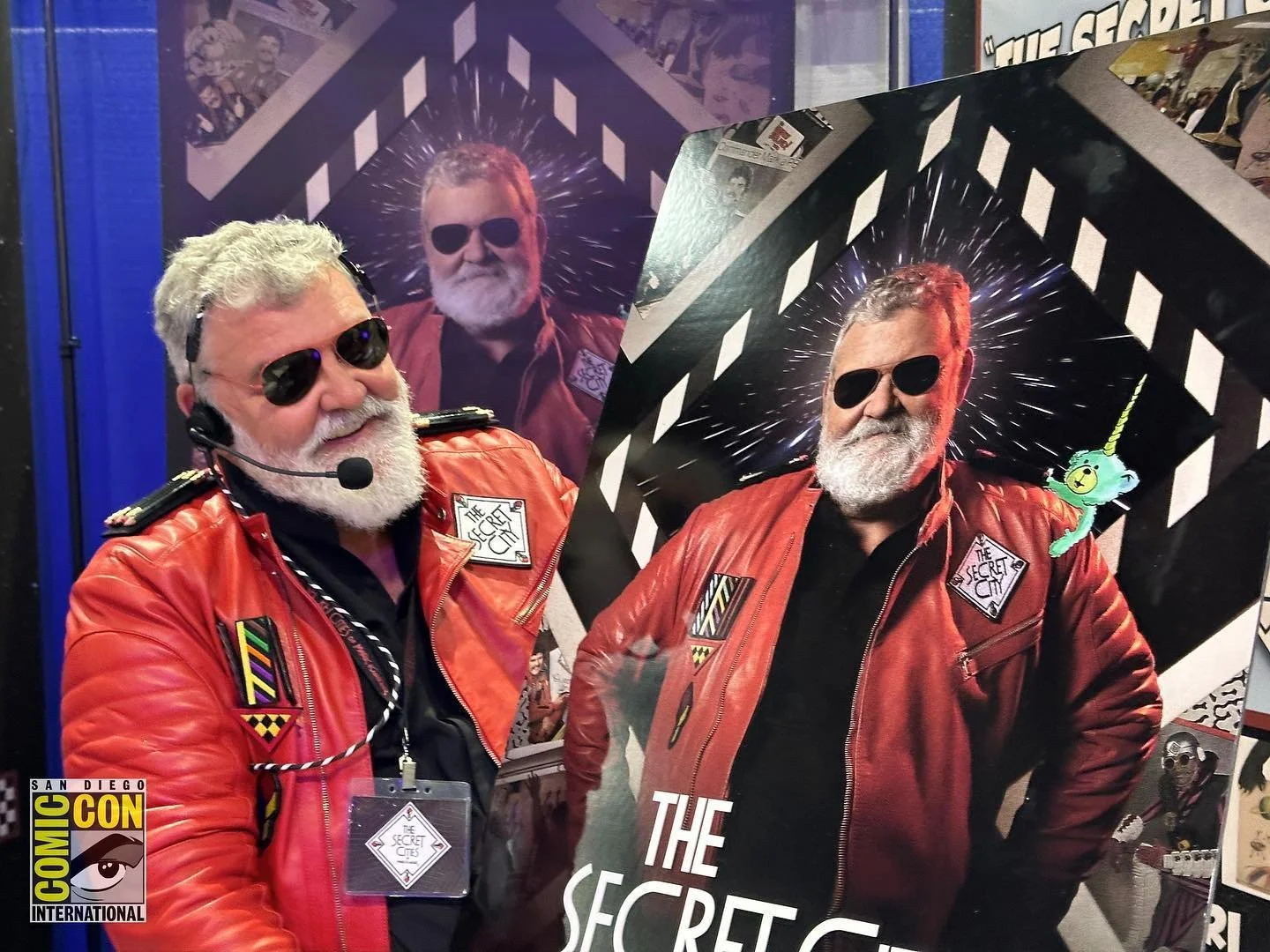 Commander Mark Kistler wearing red jacket and sunglasses, standing in front of a large poster of himself at San Diego Comic-Con. The poster features the same man in sunglasses with a cosmic background and a toy uni-bear on his shoulder.