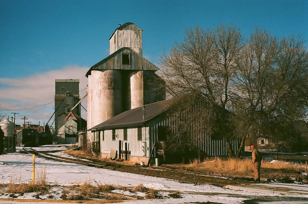 An old grain silo and a small building near railway tracks, with a person standing nearby, leafless trees, and a clear blue sky. Film photography by Jason Corgan Brown. 