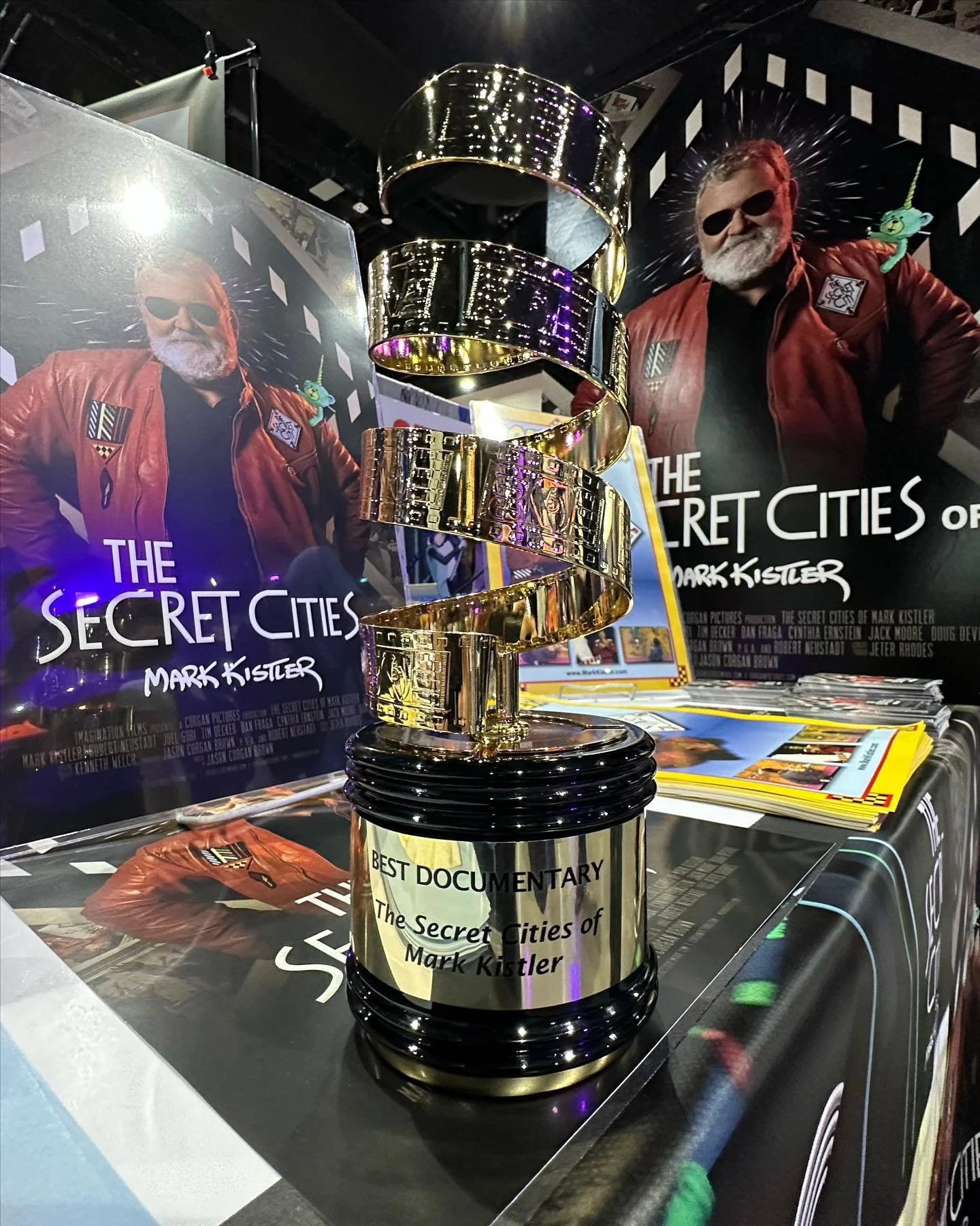 A metallic award trophy with a spiral design on top, labeled as 'Best Documentary, The Secret Cities of Mark Kistler,' placed on a table at a promotional event for the documentary.