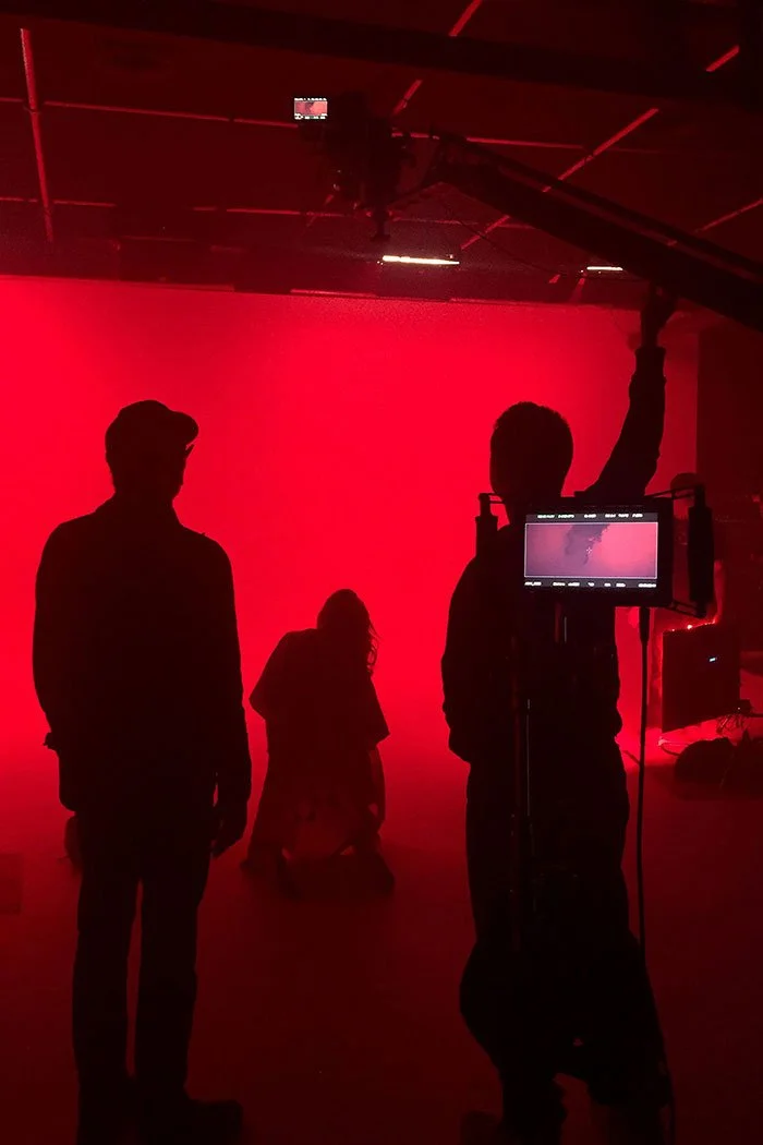Silhouettes of three people on a set with a red backdrop, one kneeling, two standing, with film equipment and a monitor visible.