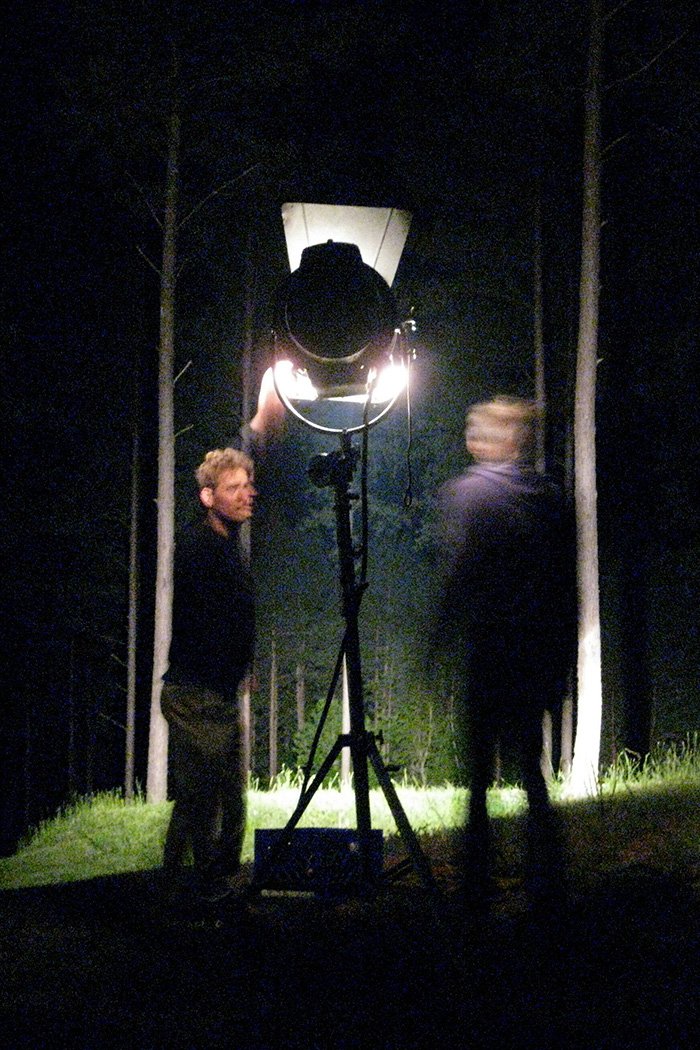 Jason Corgan Brown and Jeter Rhodes stand near a large film light in a dark outdoor setting with trees in the background, illuminated by the light. Corgan Studio film set. 