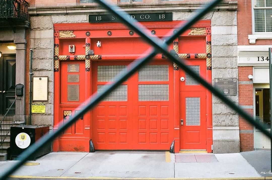 A bright red fire station garage door with decorative gold and black accents, situated between two brick buildings, partially obstructed by a black fence in the foreground. Film photography by Jason Corgan Brown. 