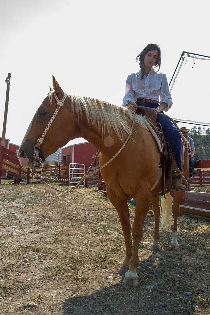 Rainey Qualley riding a tan horse with a white mane at an outdoor farm or ranch with a red barn and other people in the background.