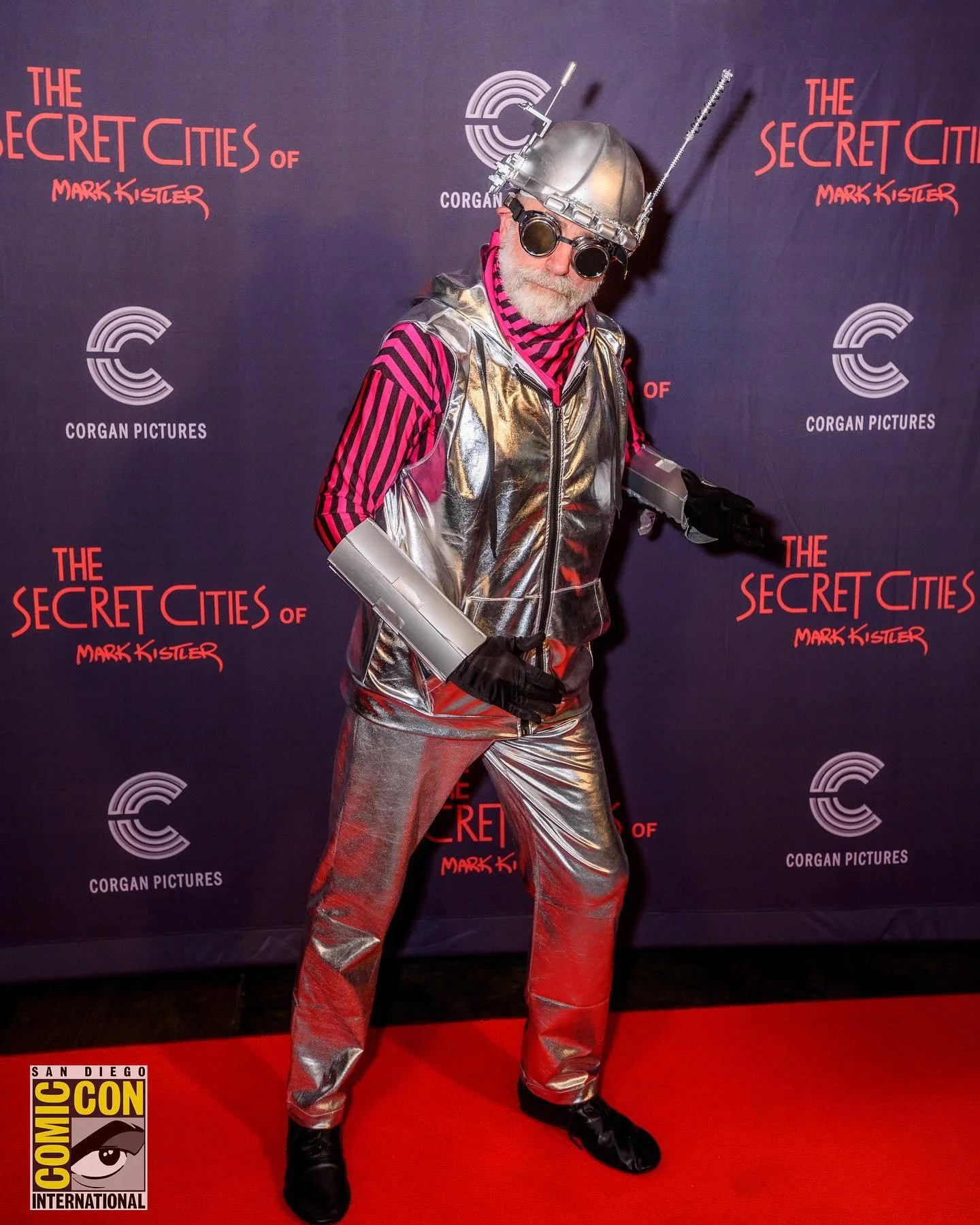 Person dressed in futuristic space-themed costume at comic convention, standing in front of a backdrop for 'The Secret Cities of Mark Kistler' by Corgan Pictures, wearing metallic silver suit, pink and black striped shirt, black gloves, black boots, 