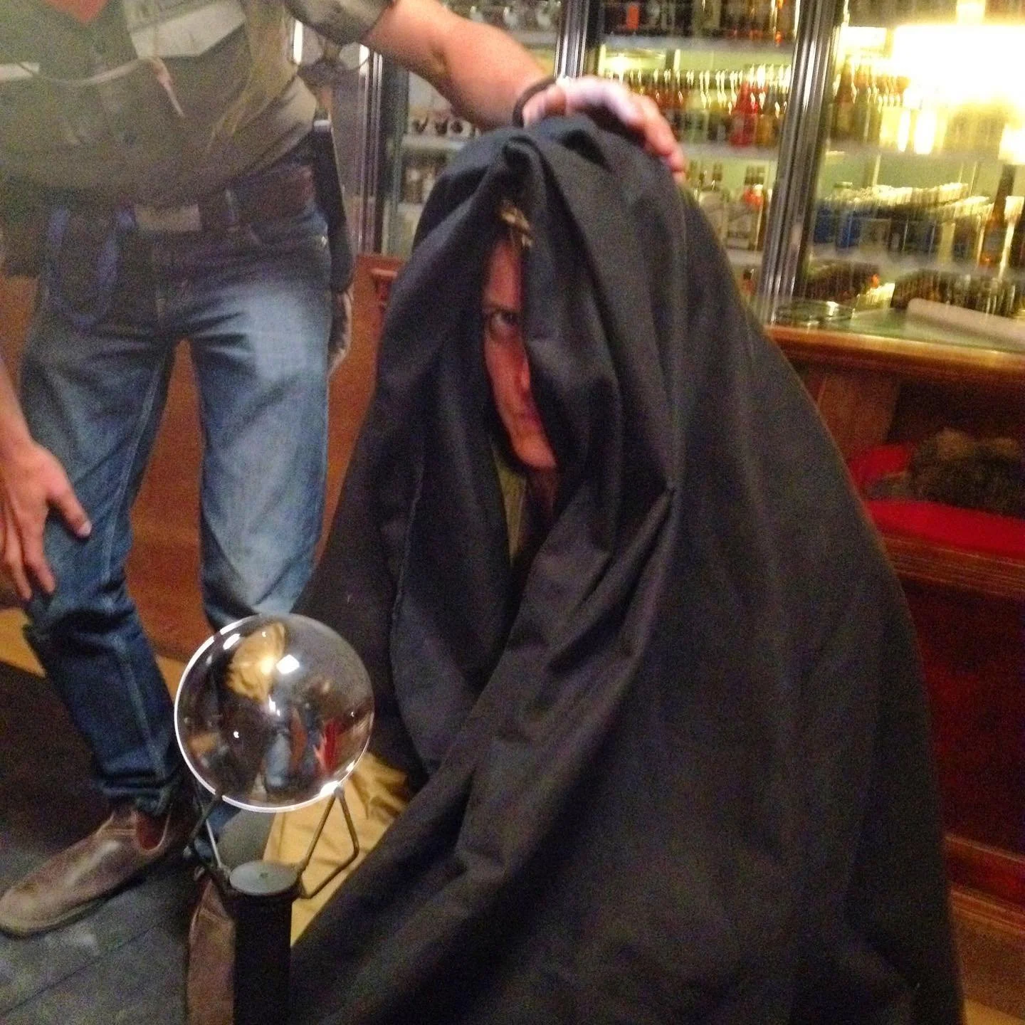 Person with a hood over their head sitting at a table in a bar or restaurant, with a small round mirror on a stand reflecting part of the person's face.