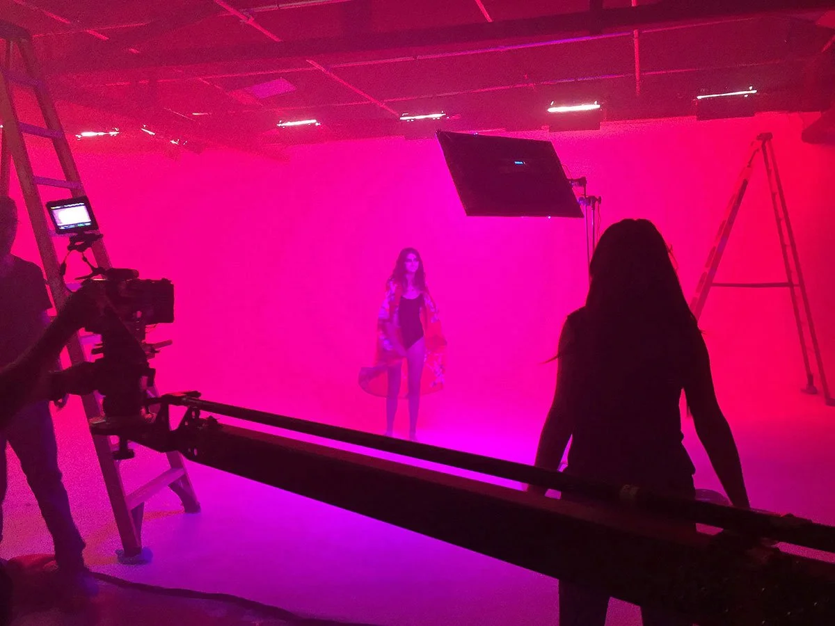 A photo of a film or photo shoot in progress with a woman model standing on a pink and purple background, surrounded by crew members, cameras, and lighting equipment in a studio.