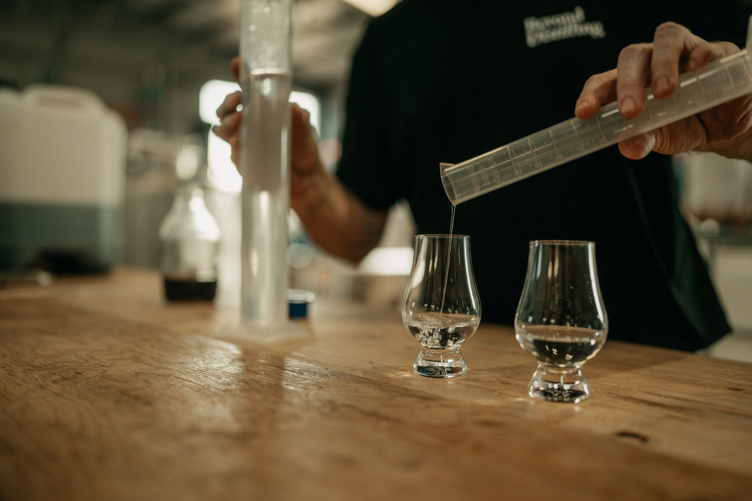 Person pouring liquid into glassware on a wooden table in a distillery or laboratory setting.