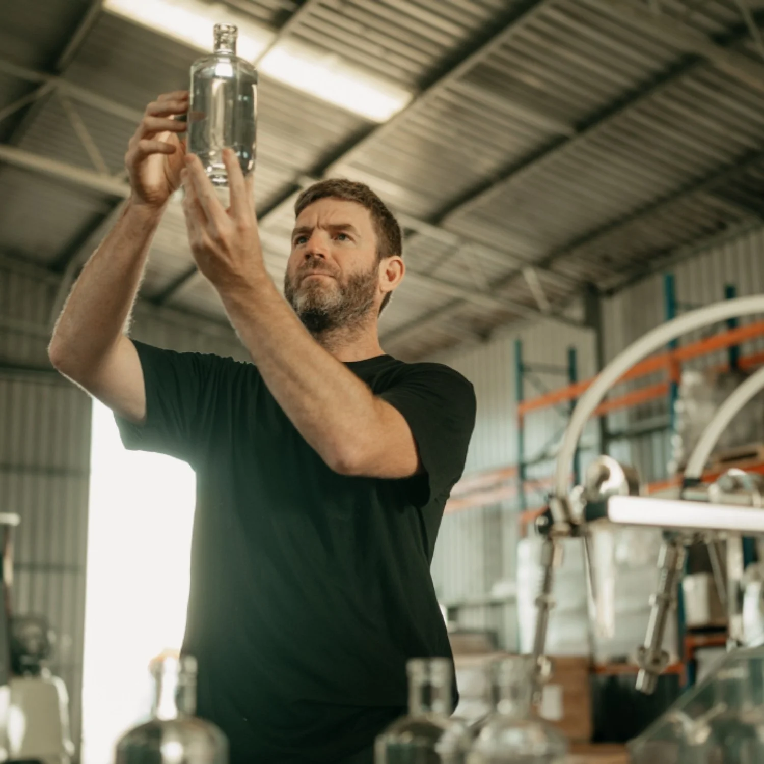 A man in a workshop or manufacturing area inspecting a clear glass bottle.