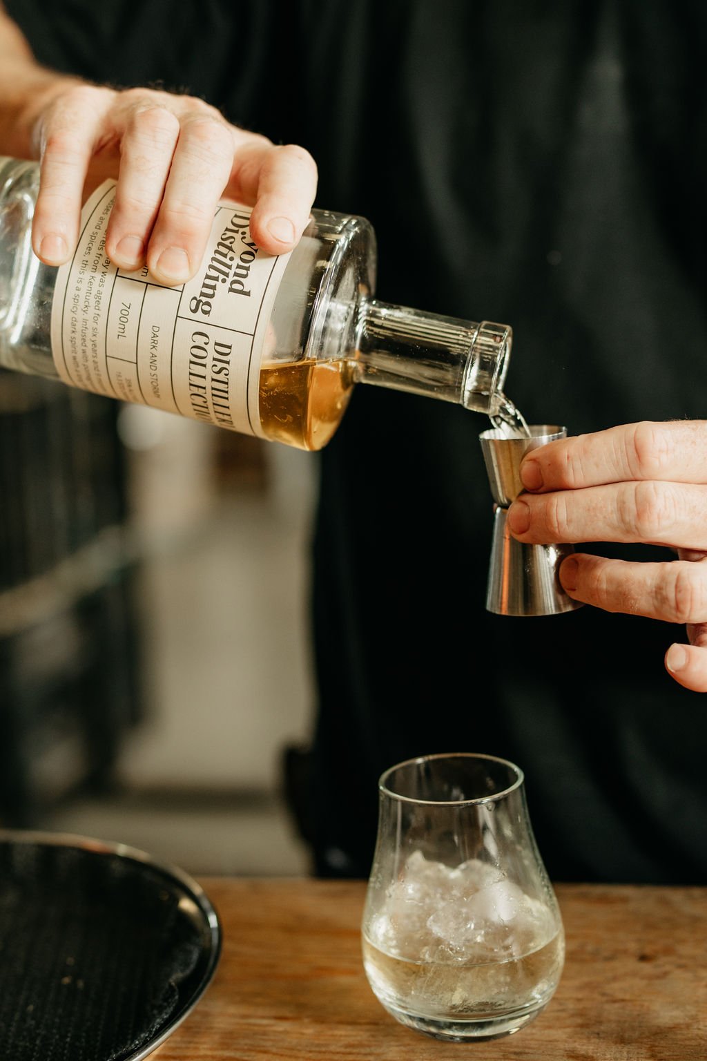Person pouring liquor from a labeled bottle into a jigger, with a glass of ice on the table.