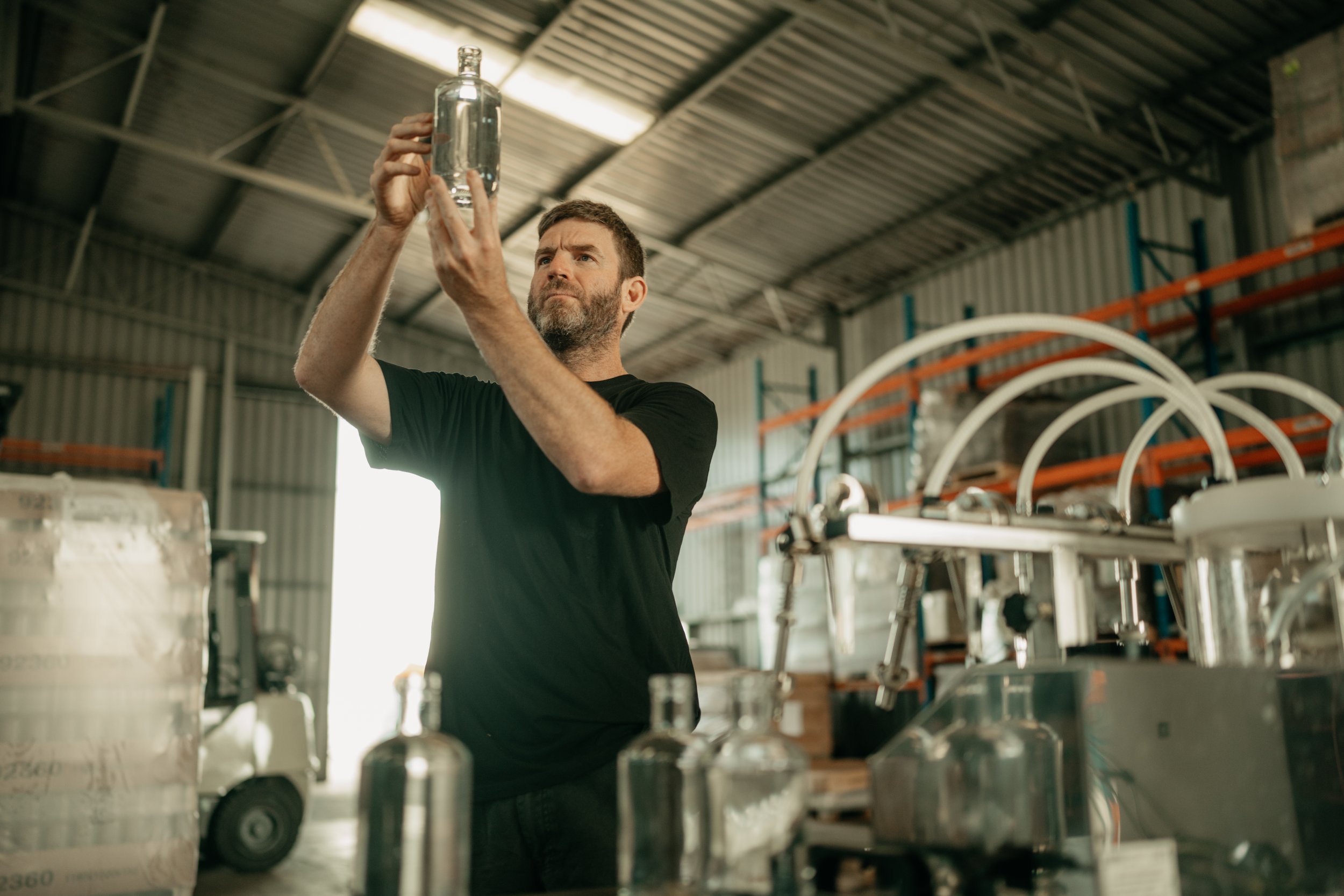 Man inspecting a glass bottle in a warehouse with filling equipment and bottles in the foreground.