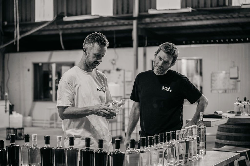 Two men in a distillery examining bottles on a table, with distillery equipment in the background.
