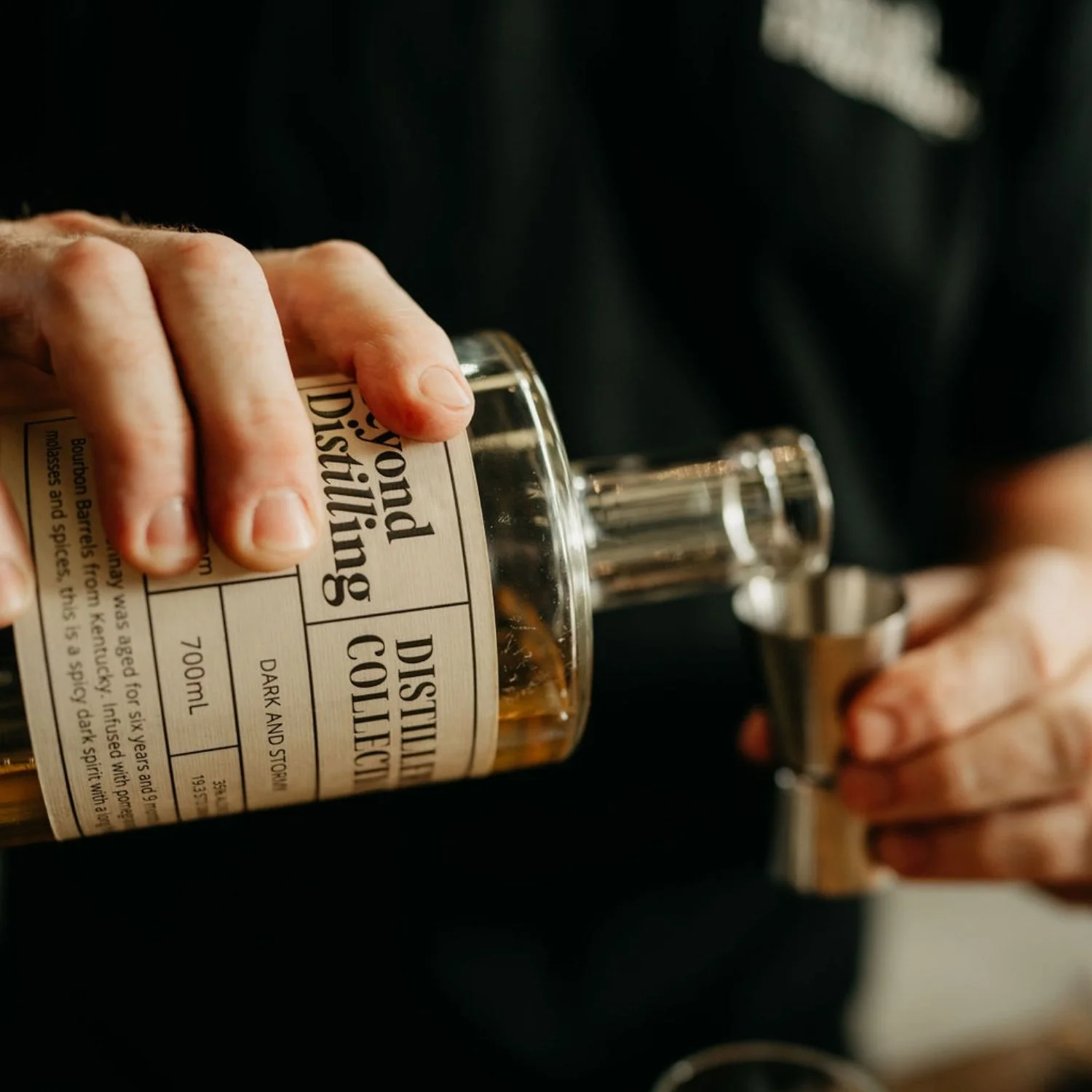 A person holding a bottle of distilled whisky in one hand and a shot glass in the other, pouring whisky into the glass.