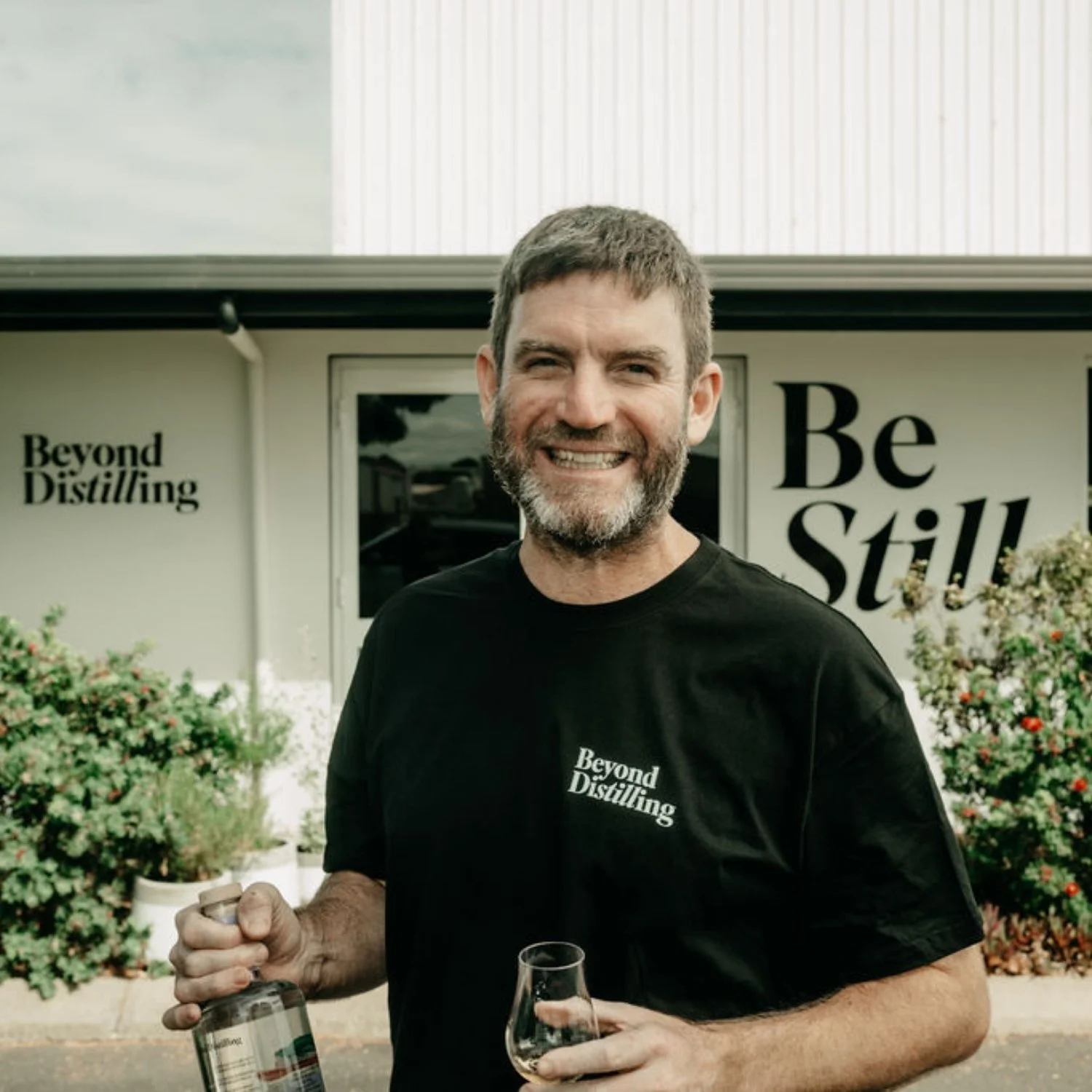 A smiling man with a beard holding a bottle and a glass of whiskey, standing outdoors in front of a building with the words "Beyond Distilling" and "Be Still" on the wall.