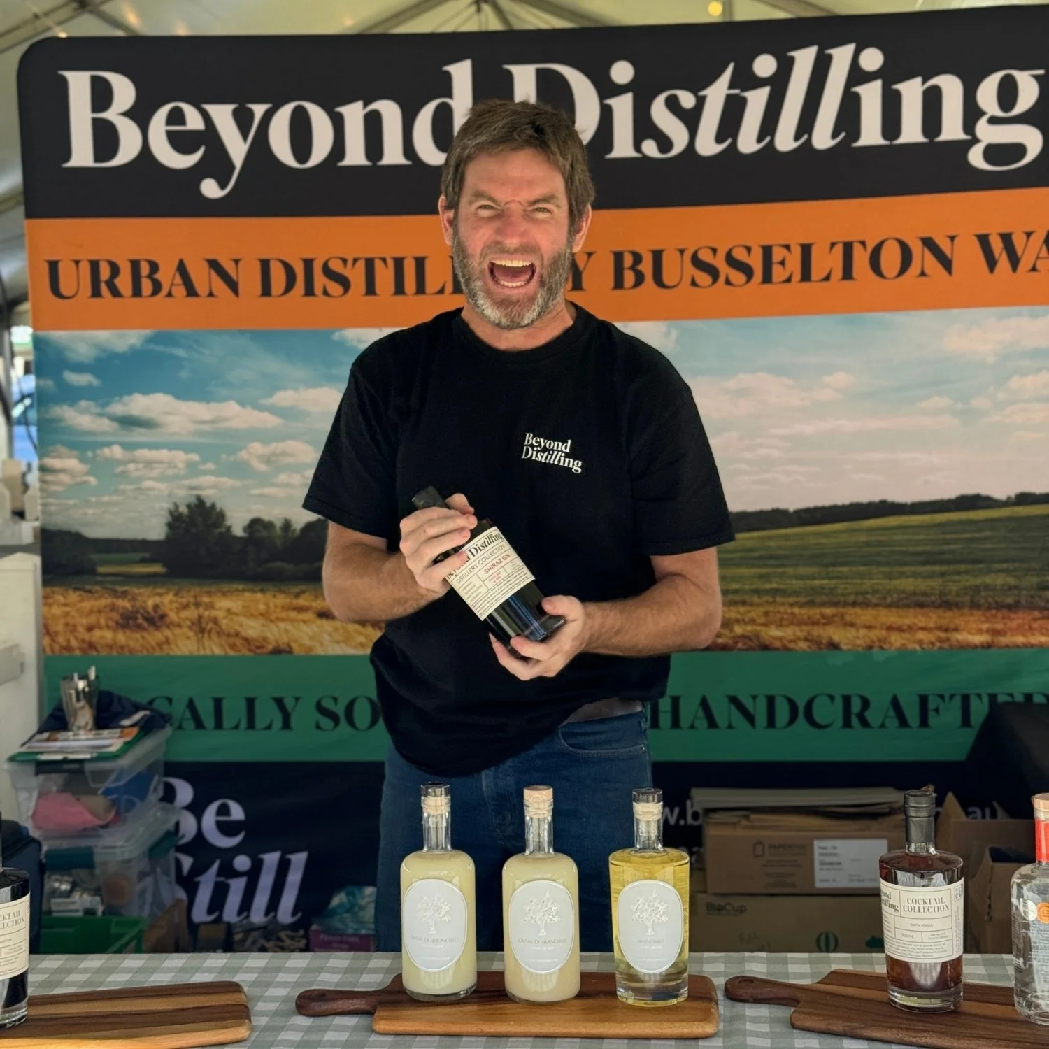 A man with a beard and gray hair, wearing a black T-shirt with white text that reads 'Beyond Distilling,' is standing behind a table displaying bottles of craft spirits. He is holding a bottle of clear liquid and appears to be talking or shouting. Be
