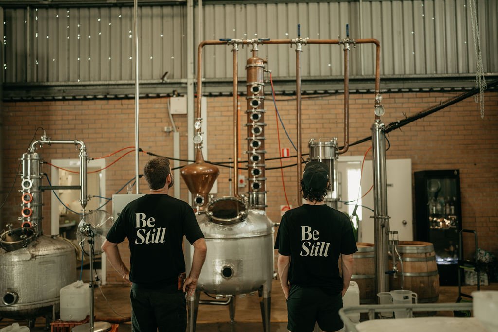 Two people in a distillery with copper and steel equipment, wearing 'Be Still' shirts.