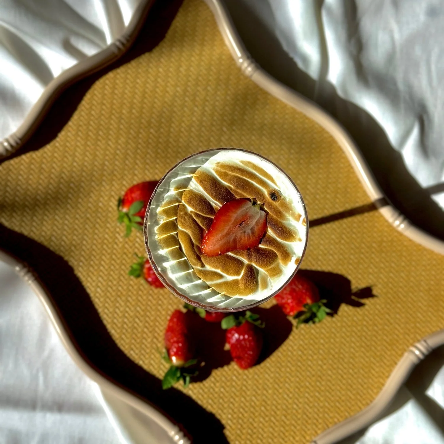 A glass of whipped cream topped with a halved strawberry on a yellow and brown patterned tray with additional strawberries.