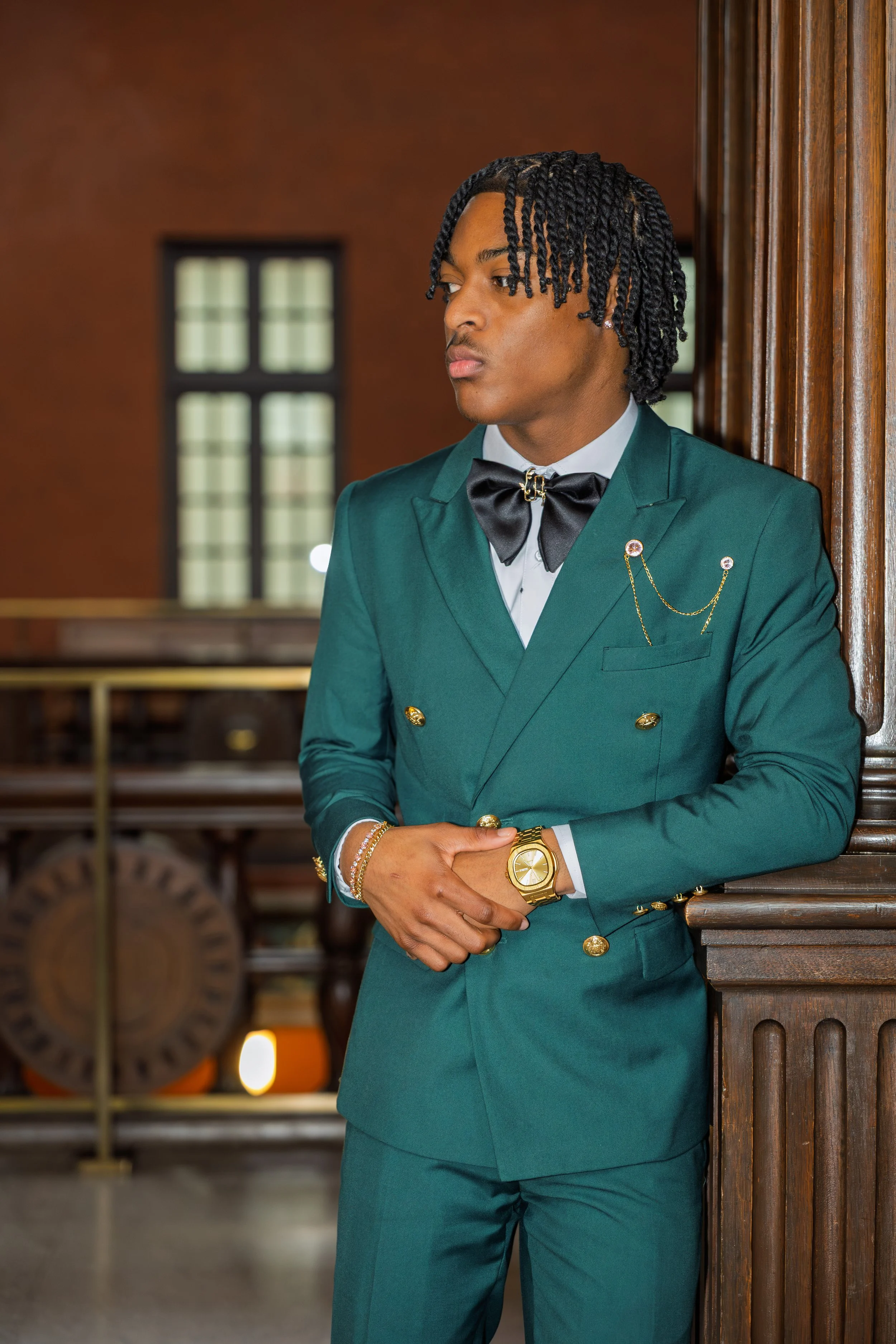 A young man in a teal double-breasted suit with gold buttons and a black bow tie stands in a wood-paneled room, leaning against a wooden column with his arms crossed and looking to the side.