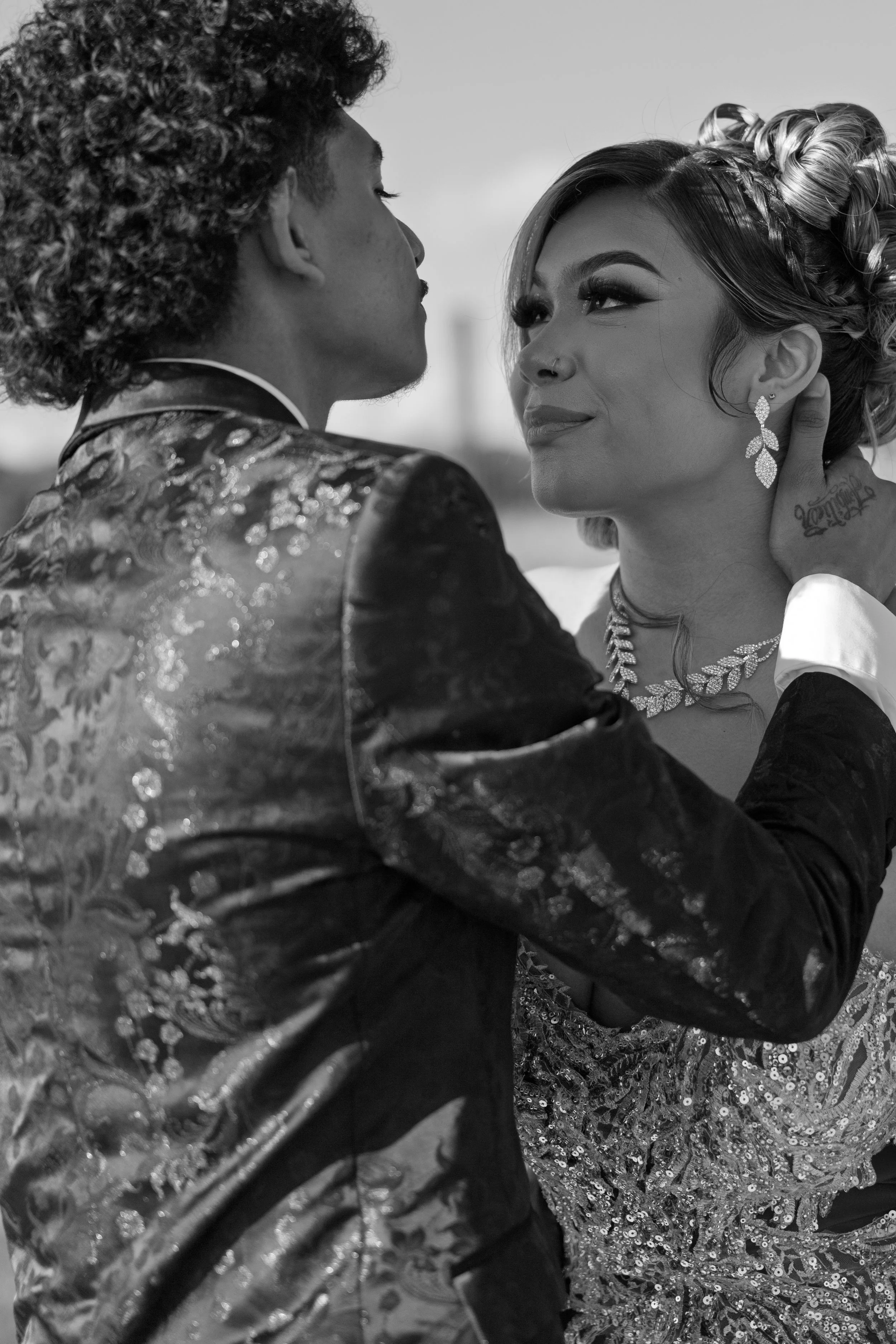A black and white photo of a couple standing close together, gazing into each other's eyes. The man has curly hair and is wearing a decorated jacket. The woman has styled hair, eye makeup, a nose ring, and is wearing earrings, a necklace, and a sequin dress. The man's hand gently touches the woman's neck.