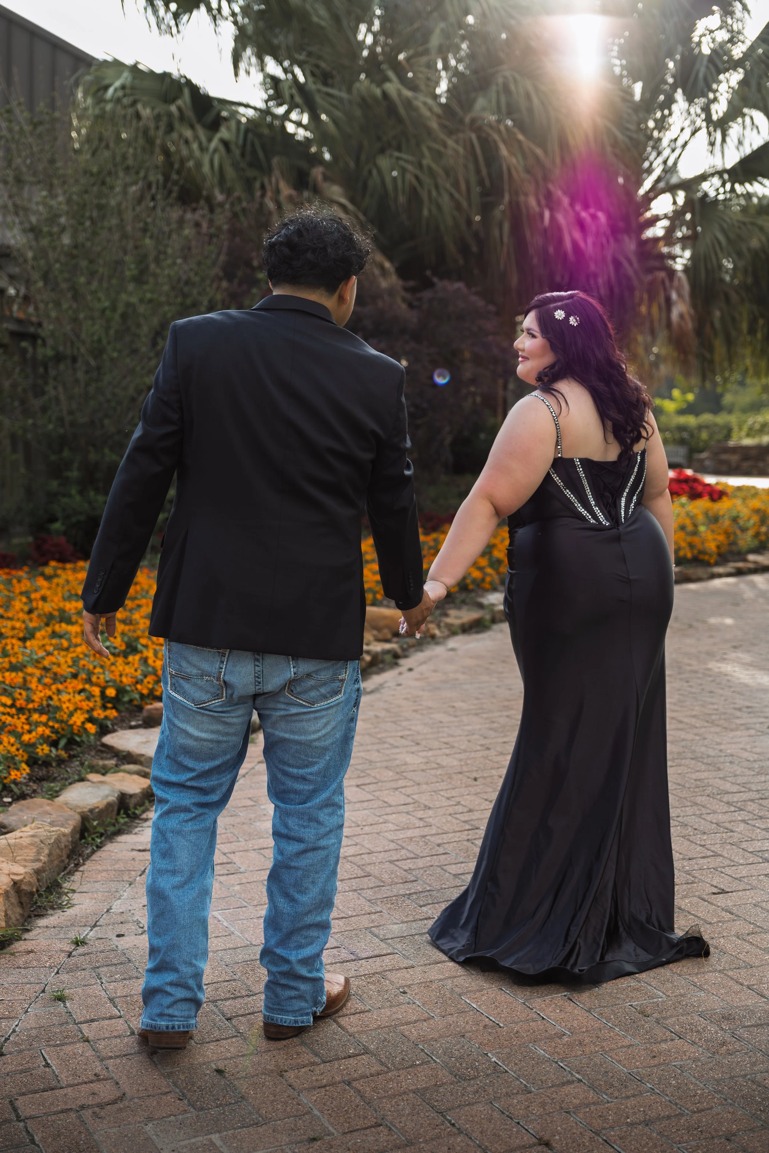 A man and a woman are holding hands while walking outdoors on a brick pathway, with the woman smiling. The man has dark curly hair and is wearing a black jacket and jeans. The woman has dark hair, is wearing a long black dress with decorative beads, and has small white flowers in her hair. The background includes trees, flowers, and sunlight.