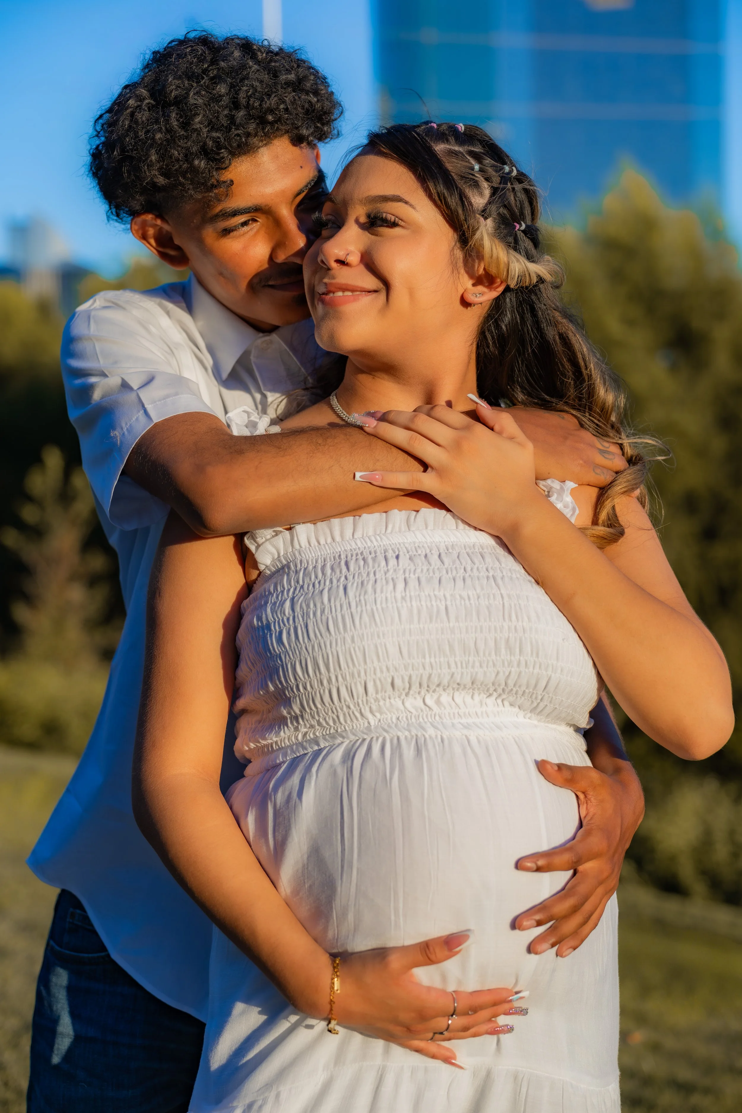 A couple hugging outdoors in the sunlight, with the woman's pregnant belly visible, both smiling warmly.