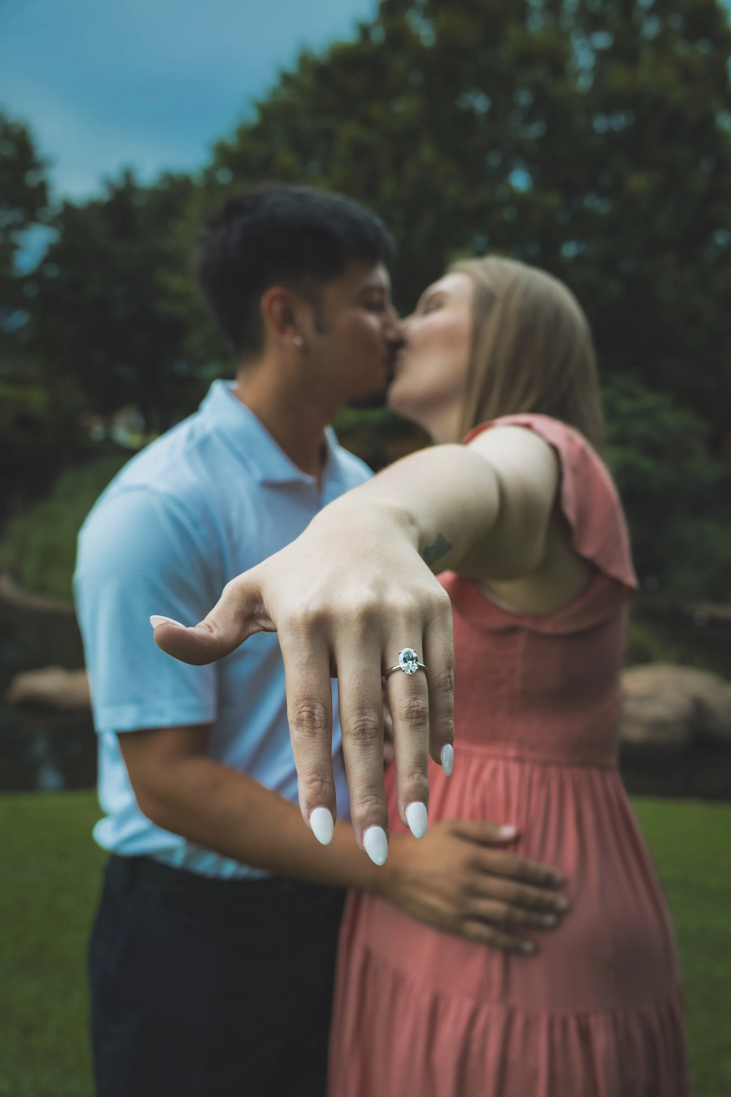 A couple kissing outdoors with a woman showing off an engagement ring on her finger, while the man is touching her pregnant belly.