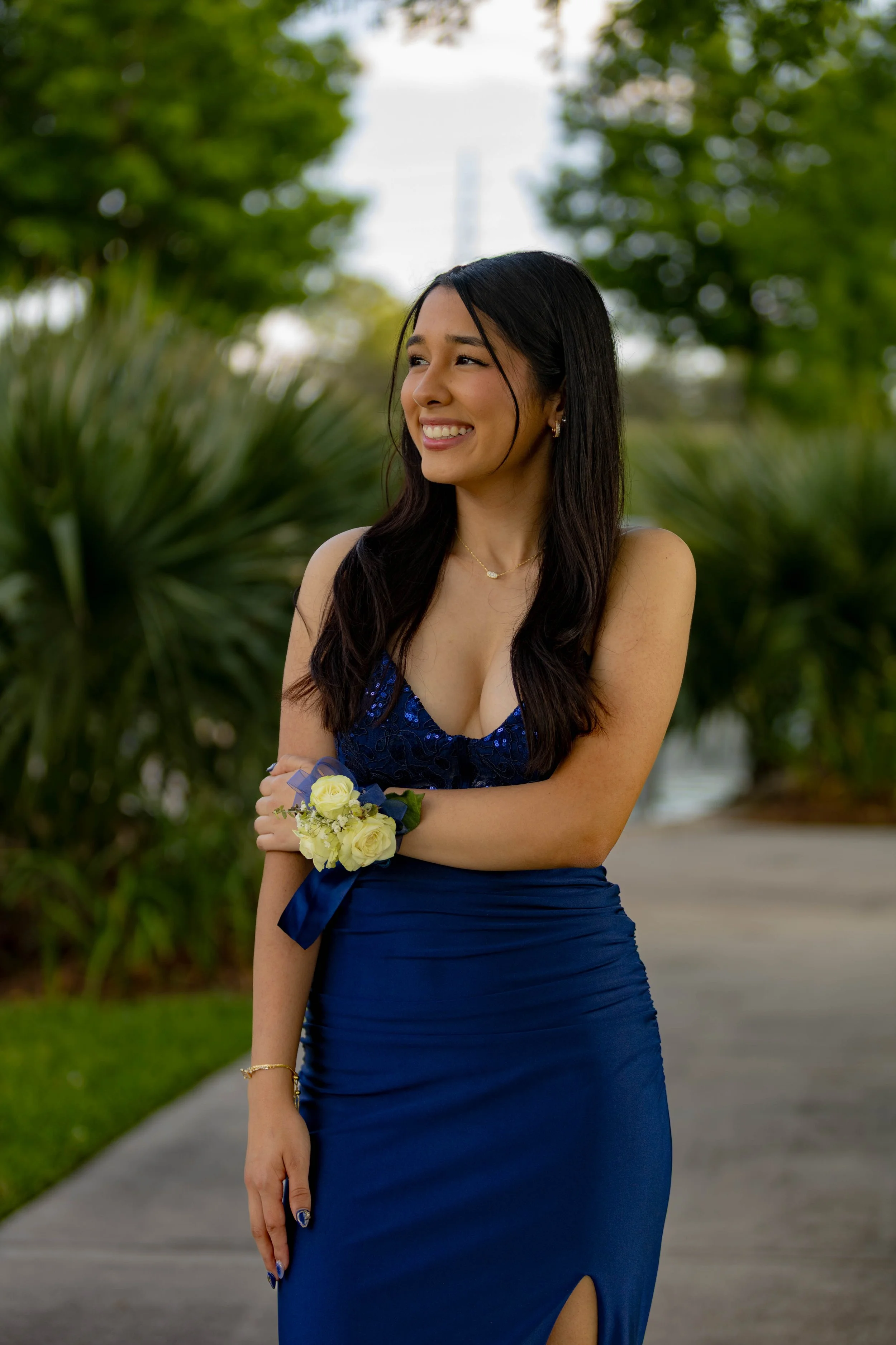 A young woman in a blue evening gown is standing outdoors, smiling, with arms crossed holding a small bouquet of yellow roses. She has long dark hair, wearing earrings, a necklace, and a bracelet, with a background of trees and greenery.