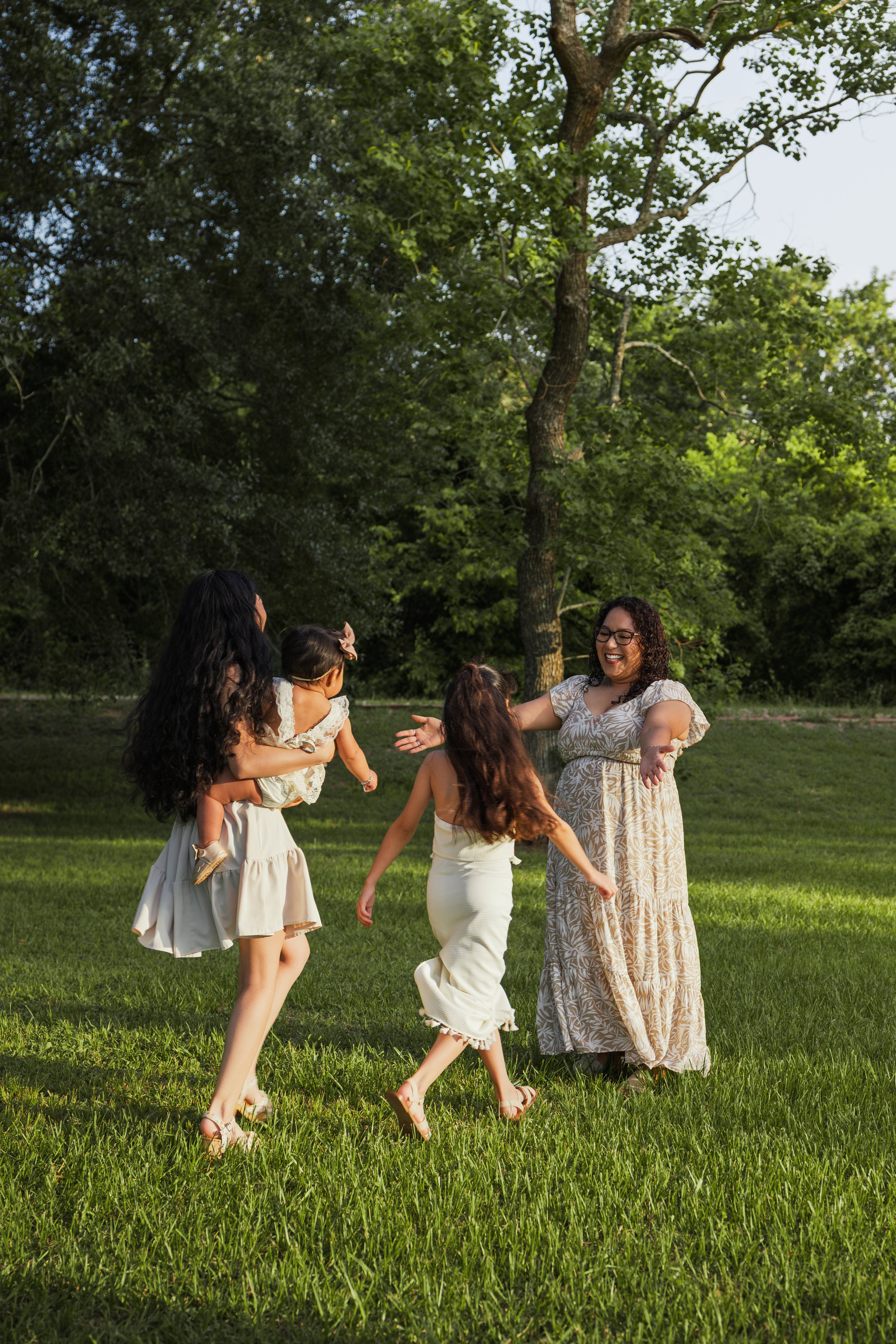 A woman in a long dress is smiling and extending her arms while a group of children, some holding hands, dance and play on a grassy field under tall trees.