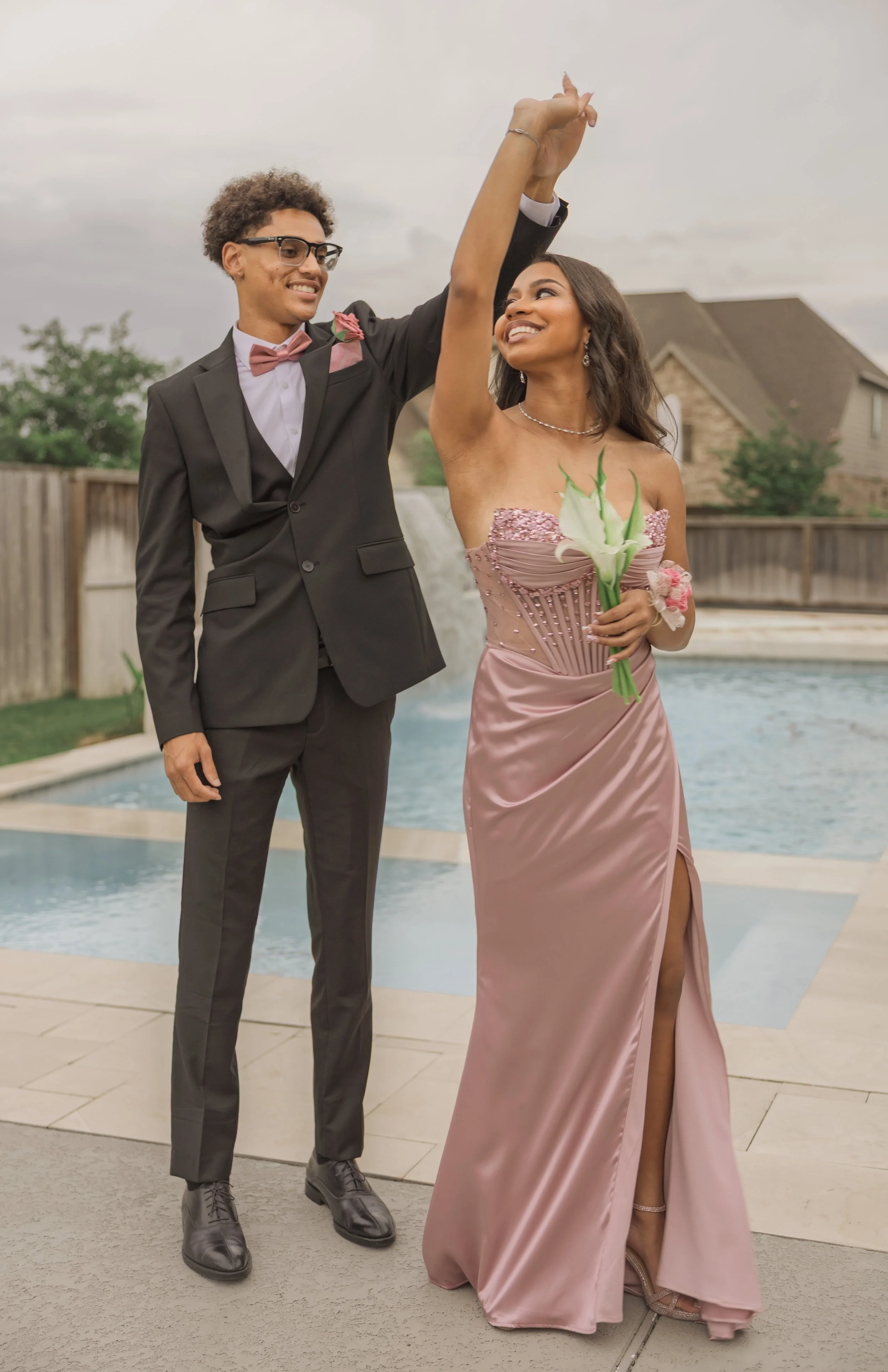 A young man and woman are dancing by a pool. The man is wearing a tuxedo with a pink bow tie and a pink rose boutonnière, and the woman is in a strapless pink gown holding a white calla lily. They are smiling, with the man raising his hand and the woman looking at him up close. There are houses and trees in the background.