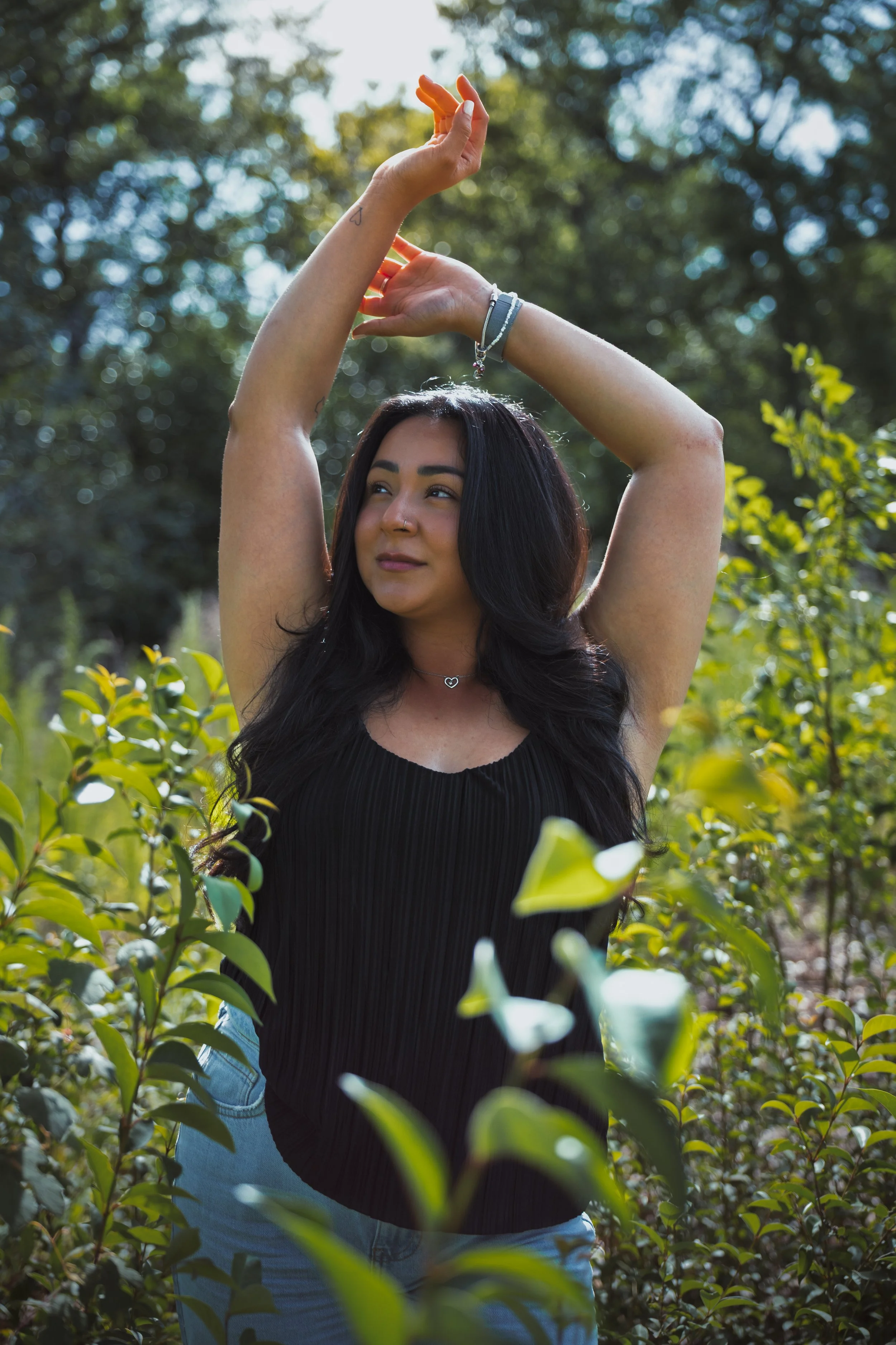 A woman with long dark hair and a nose ring standing among green leafy plants, raising her arms above her head with her eyes closed and a slight smile.