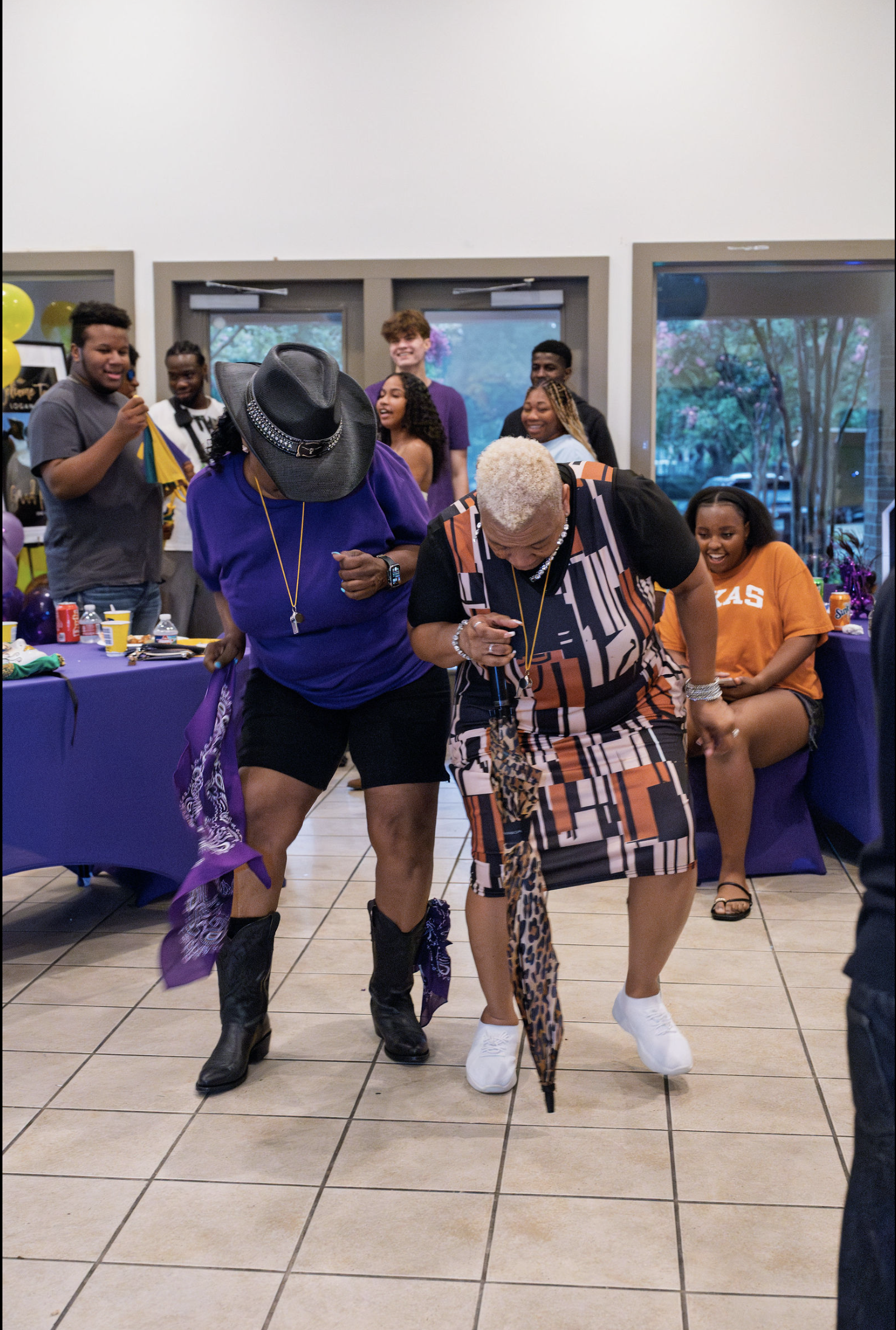 Group of people at a party or celebration, dancing and laughing, with a table of snacks and balloons in the background.