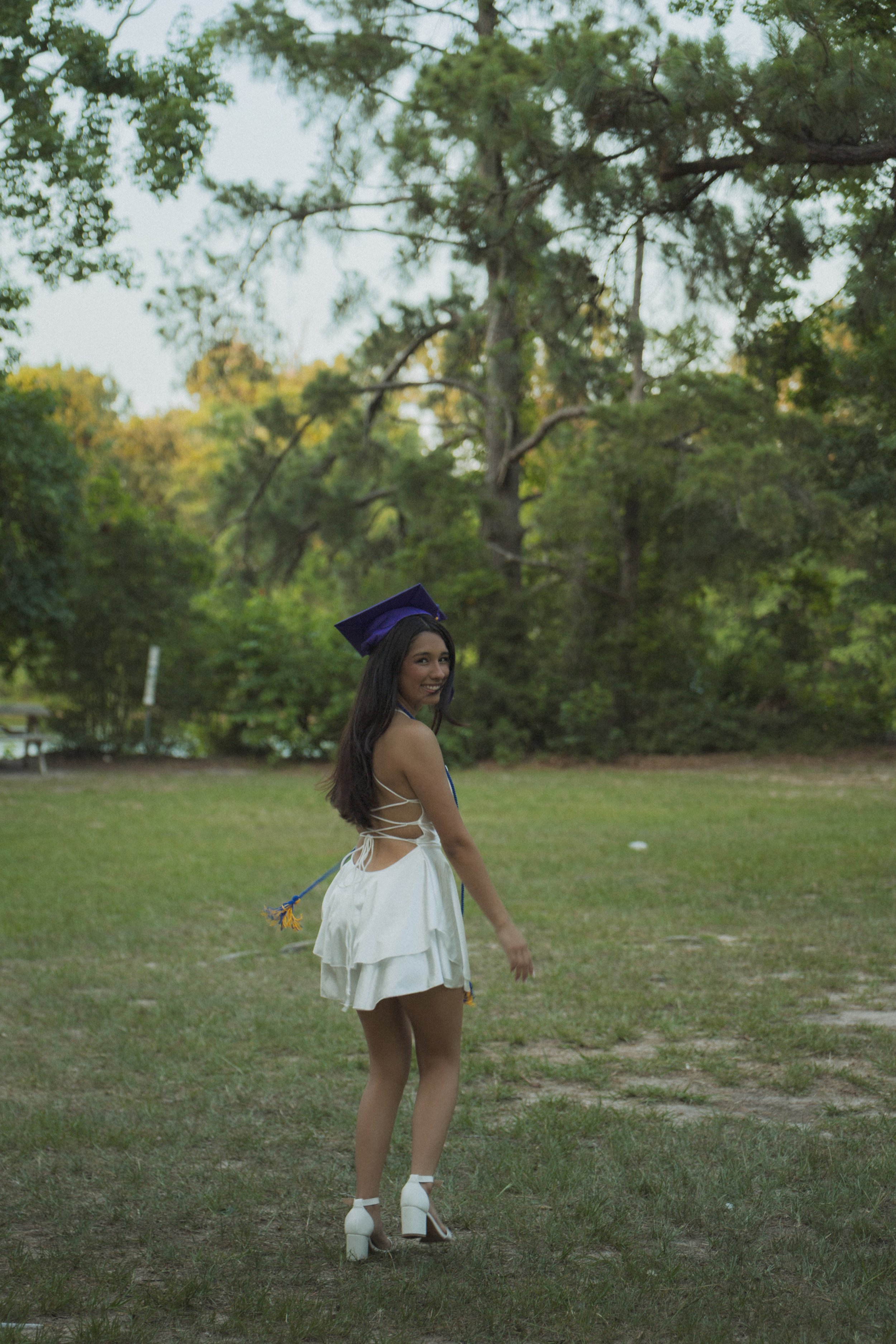 Young woman in a white dress and high heels standing outdoors on grass with trees in the background, smiling, wearing a purple graduation cap.