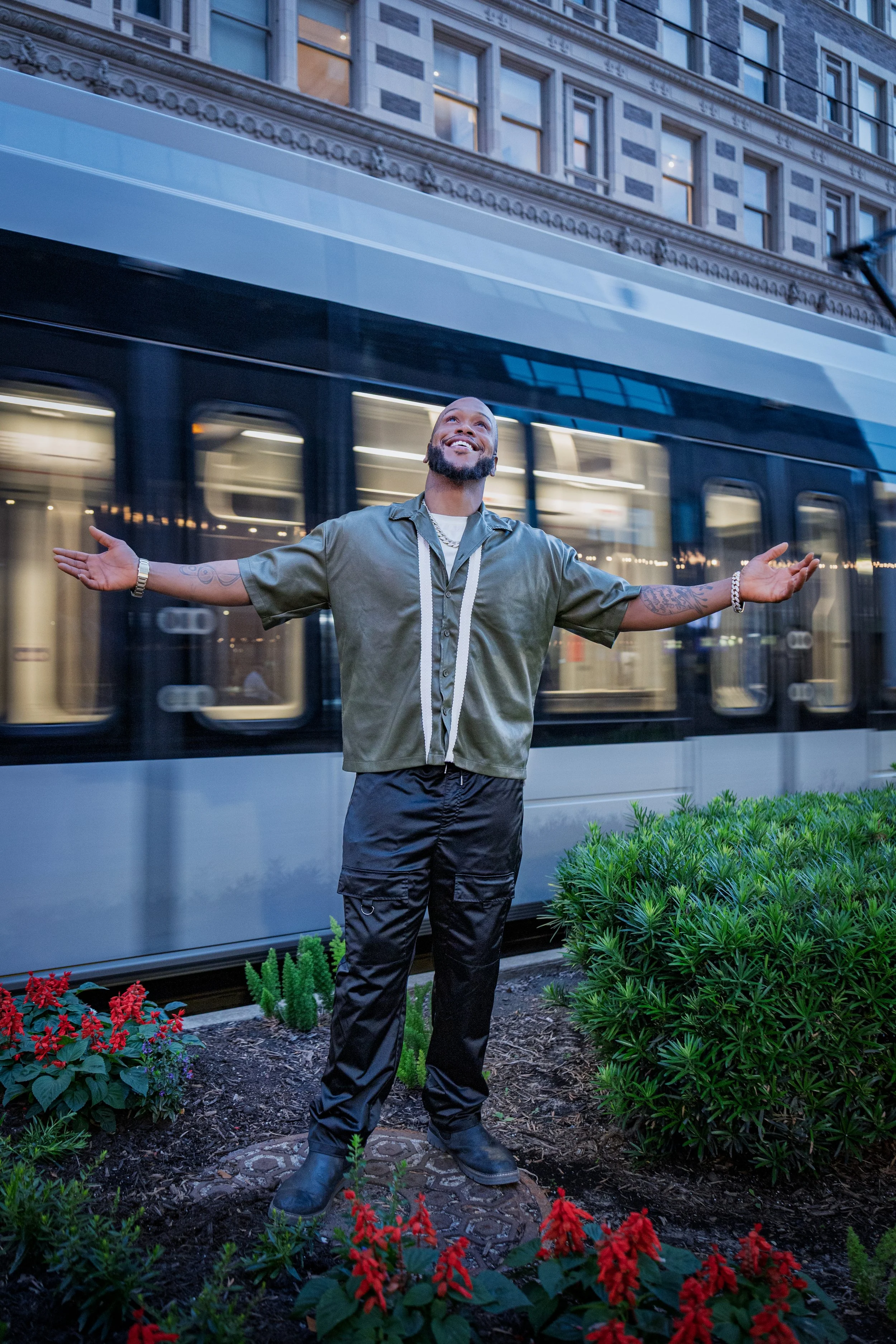 A man with arms outstretched stands in front of a city train with a historic building in the background, surrounded by flowers and greenery.