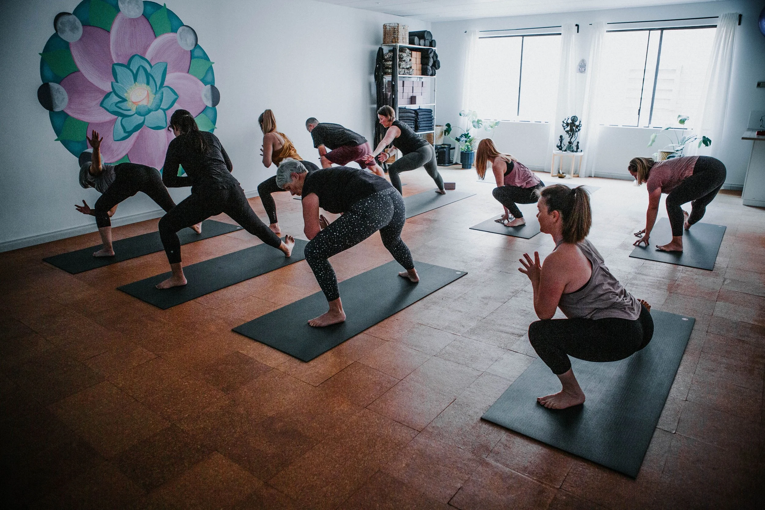 A group of diverse people participating in a Bloom yoga class in a bright, spacious room with large windows, yoga mats, and a lotus moon mandala mural on the wall in Kingsley, Perth