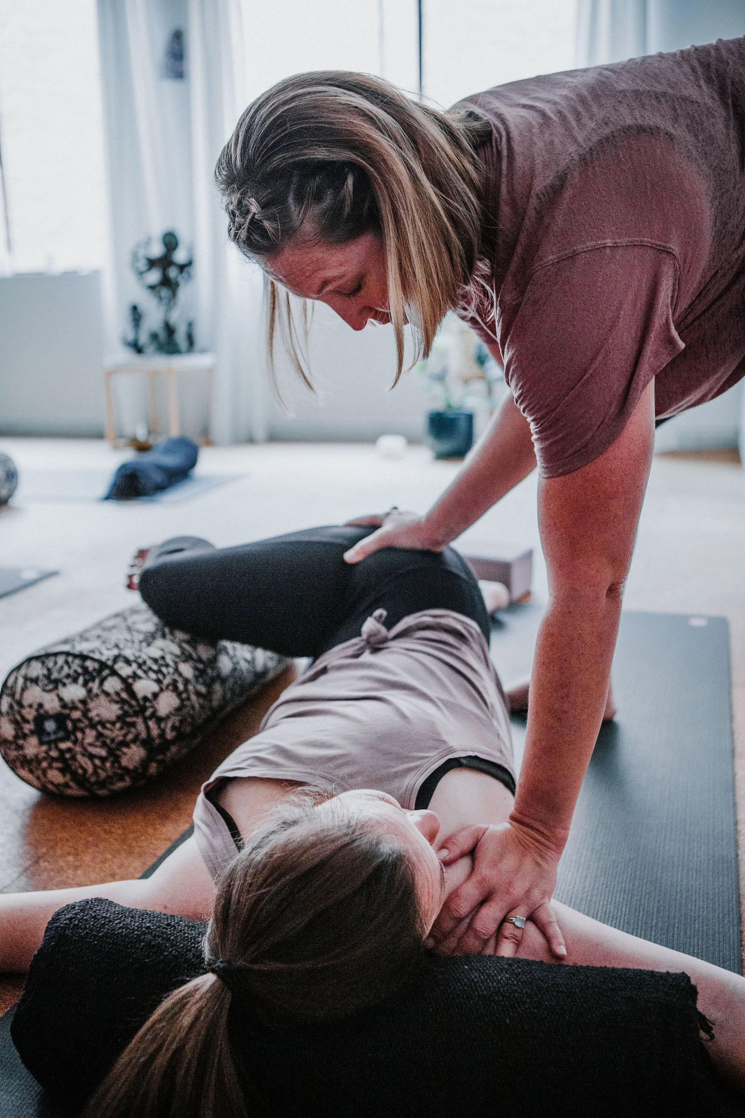 A woman receiving a yoga assist during a restorative yoga pose while lying on a yoga mat in a light, bright Kingsley yoga studio.
