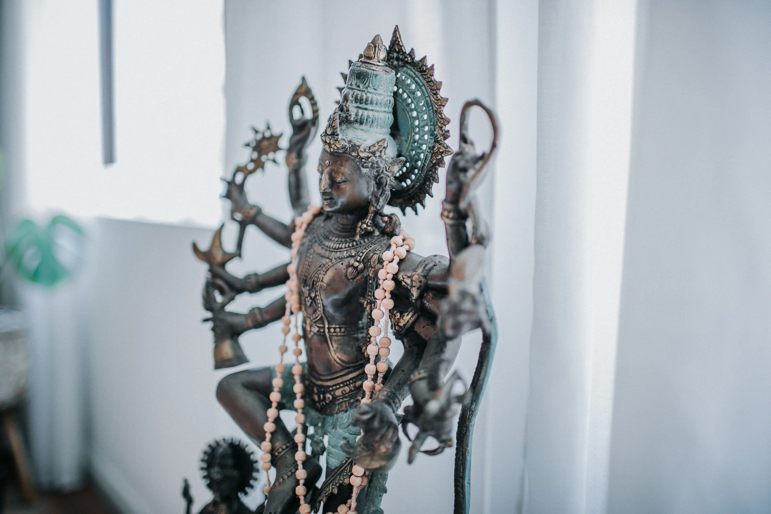 A bronze-colored statue of a multi-armed Hindu deity, adorned with necklaces and an elaborate headdress. The statue is set against a light background, with some blurred green plant in the foreground.