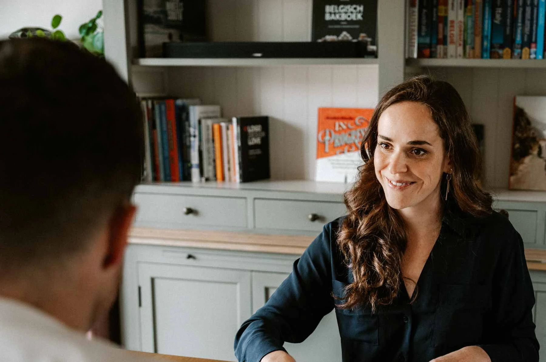 Een vrouw met lange, golvende bruine haar glimlacht terwijl ze met een man spreekt in een interieur met boekenplanken vol boeken op de achtergrond.
