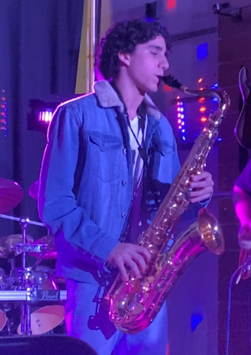 Young man playing the saxophone in a dimly lit environment with colorful lights.